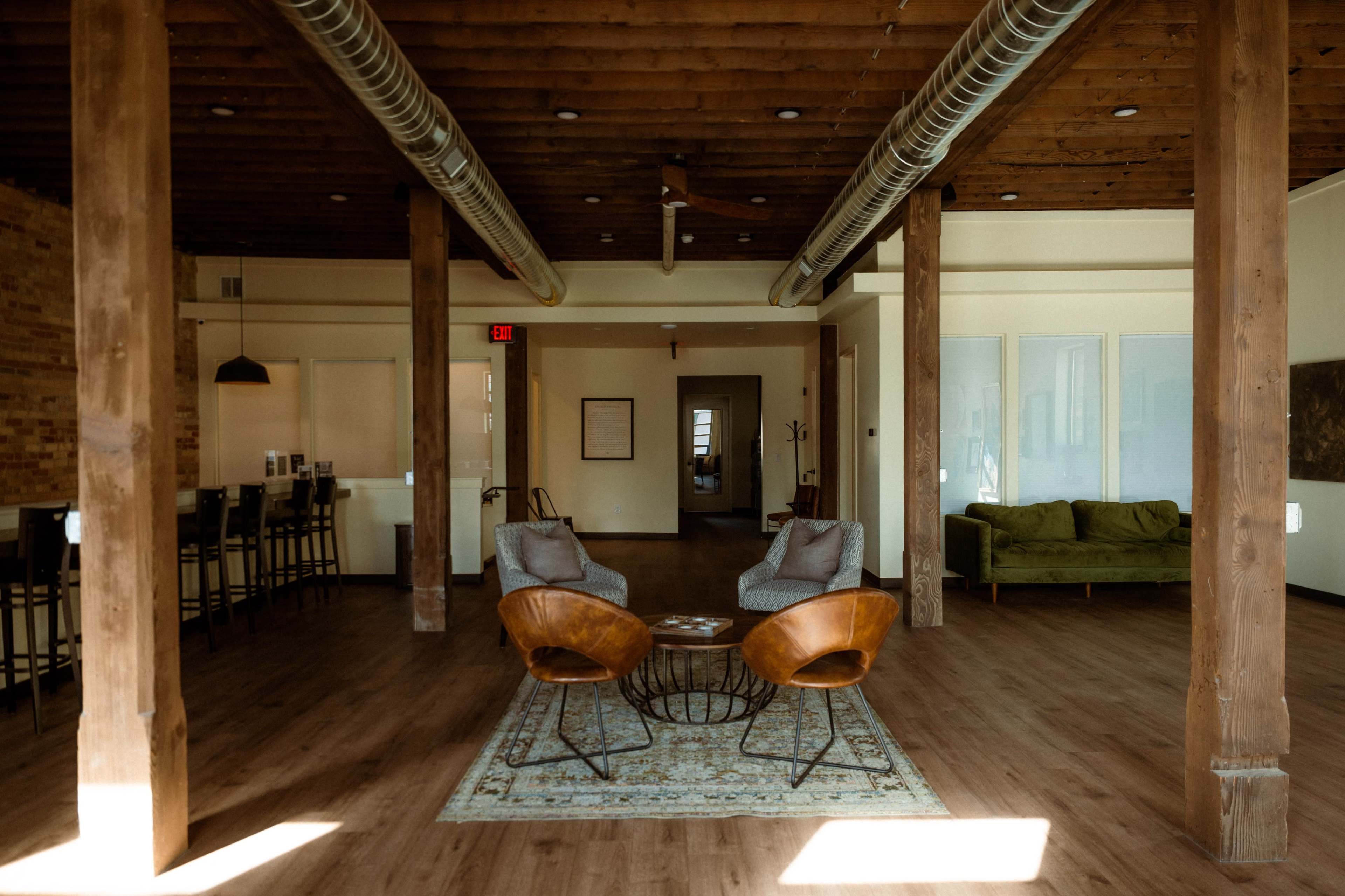 The image shows a spacious, modern interior with wooden beams, a seating area featuring two chairs and a round table, and an open layout leading to different rooms.