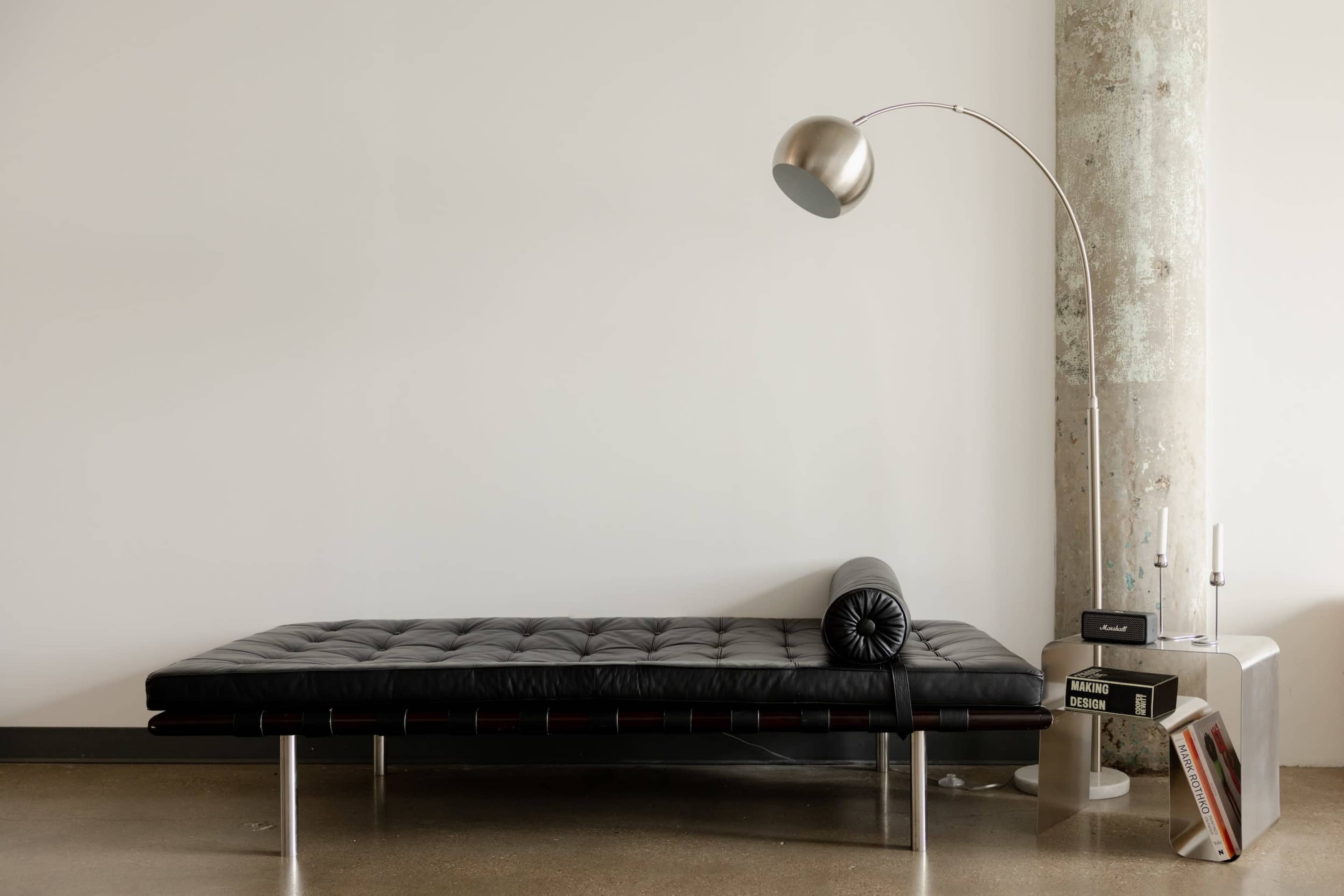 The image shows a minimalist black leather daybed next to a curved floor lamp and a small side table with a stack of books.