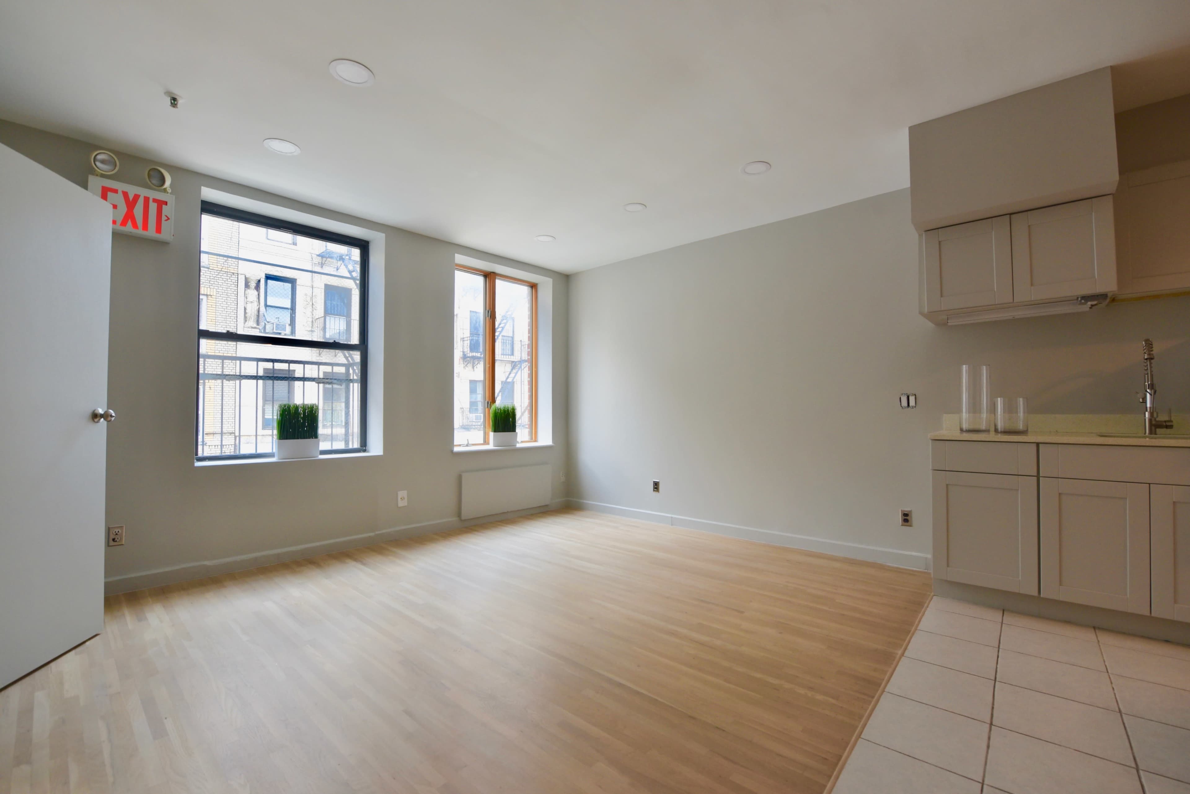 A spacious, empty room with two large windows and a kitchen area on the right.