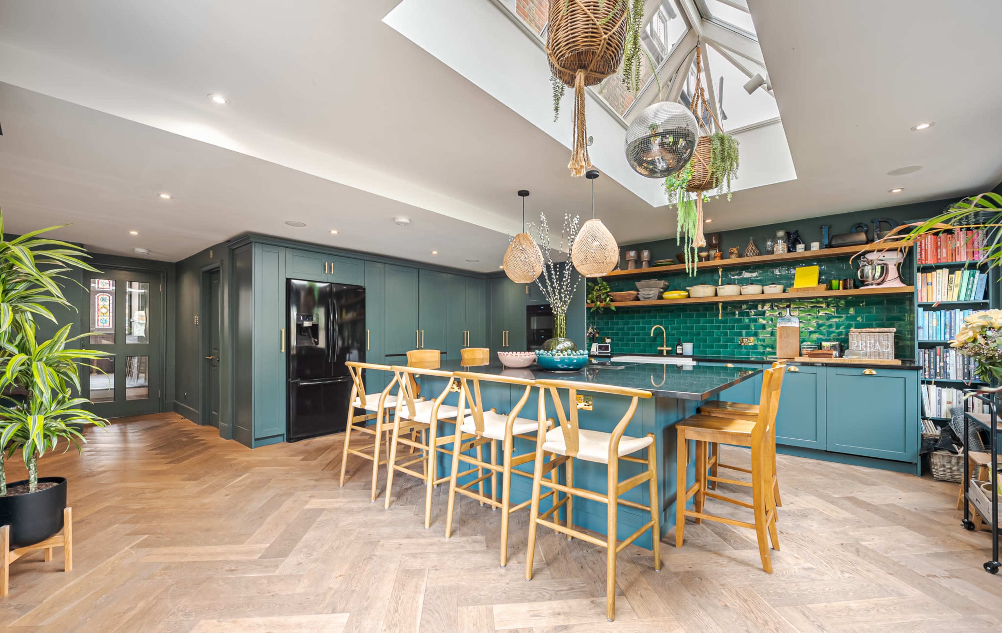 A modern kitchen features a large island with wooden bar stools, green tile backsplash, and a skylight overhead, surrounded by plants and shelves.