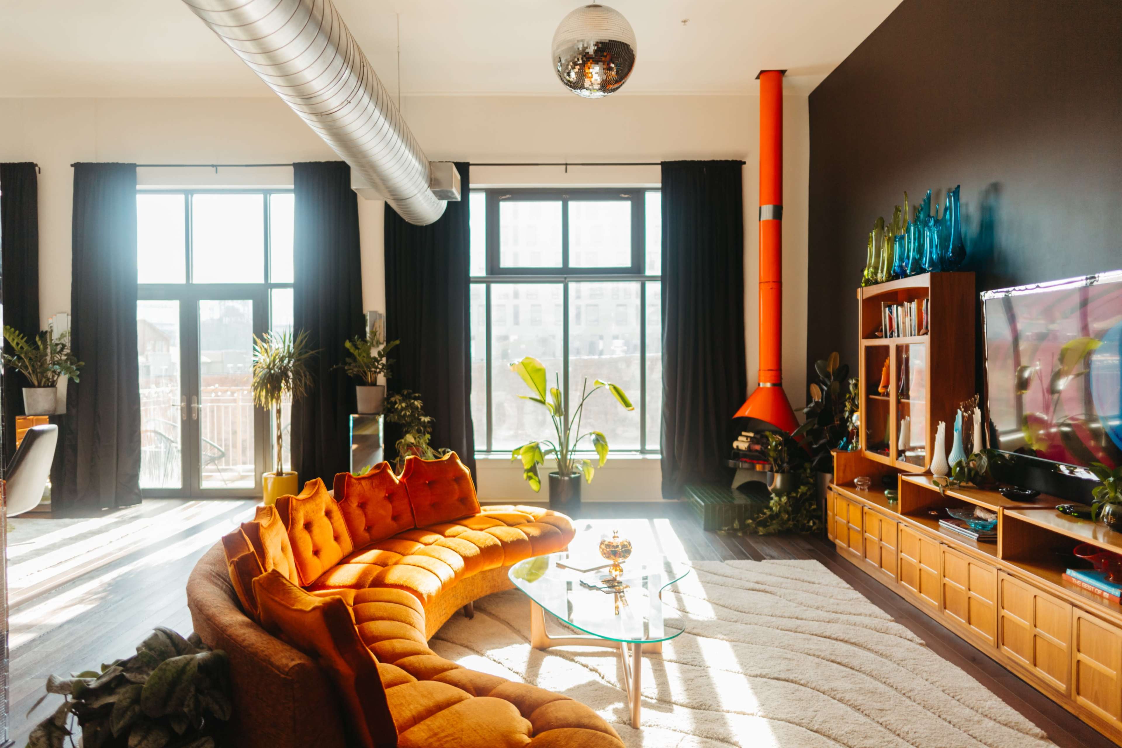 A bright, modern living room features a curved orange sofa, a glass coffee table, large windows with sheer curtains, and a colorful fireplace.