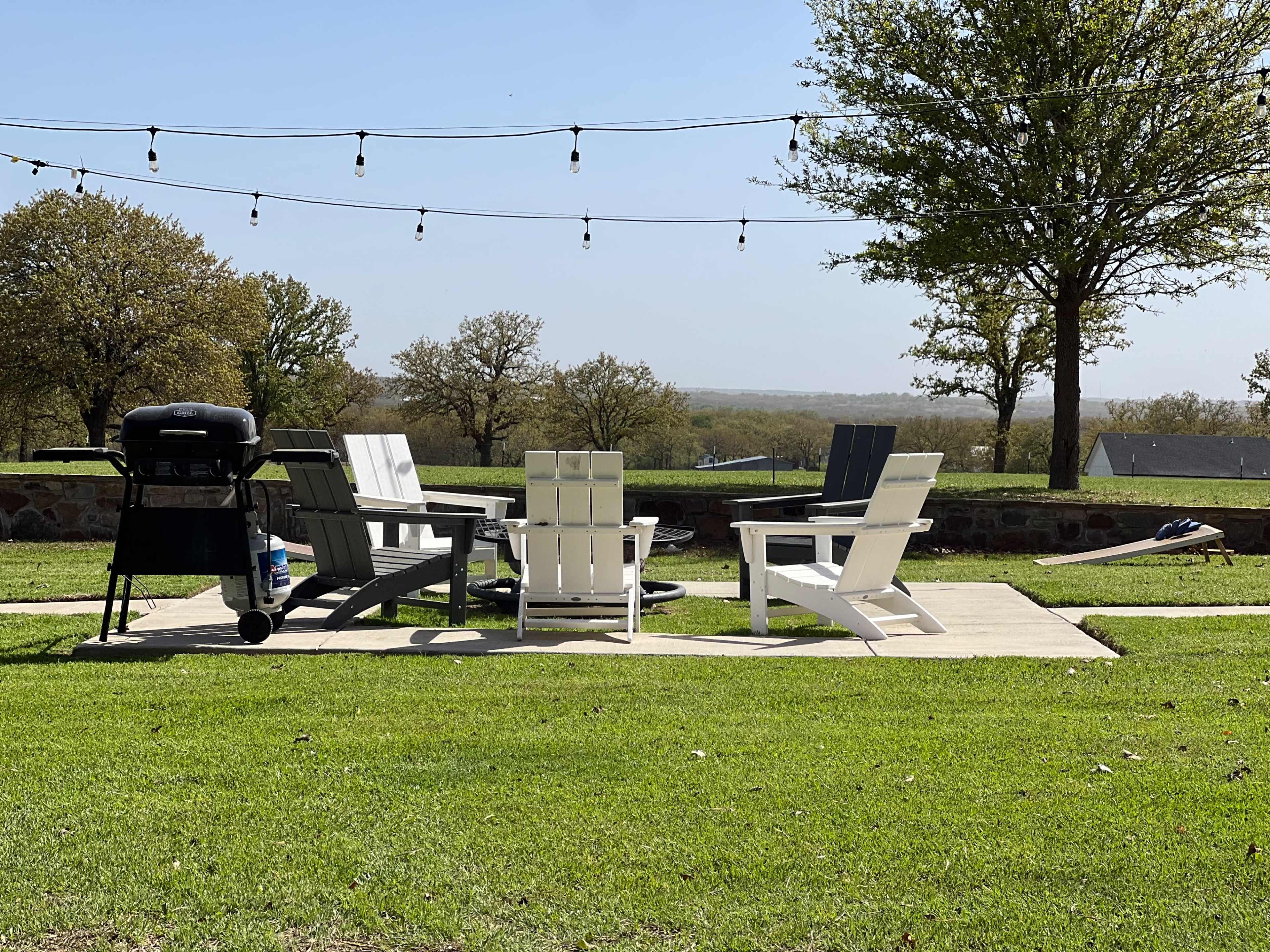 The image shows a patio area with four lawn chairs and a grill, surrounded by green grass and trees, under string lights.