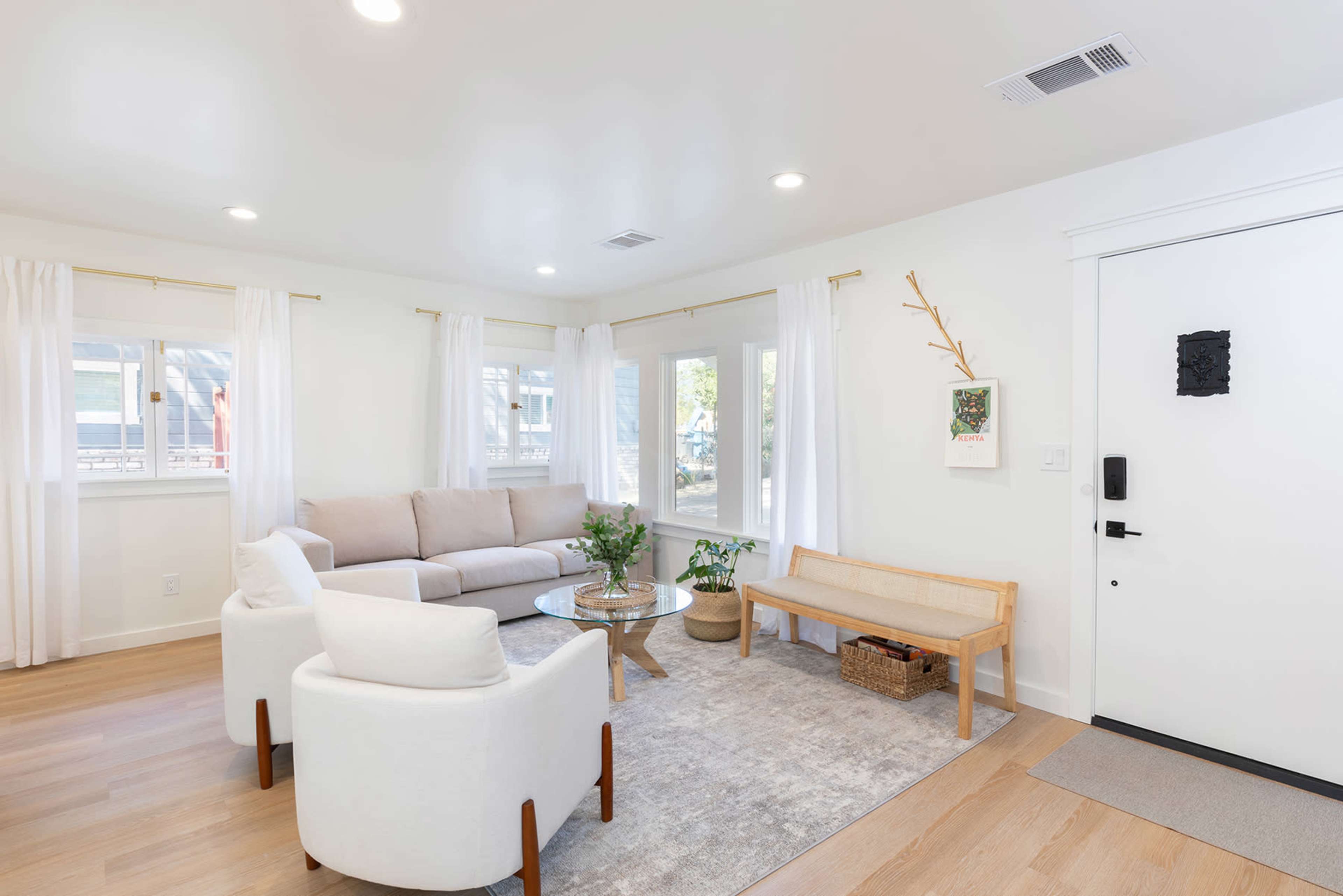 A bright living room featuring a light-colored sofa, armchairs, a coffee table, and a decorative bench against a white wall with large windows.