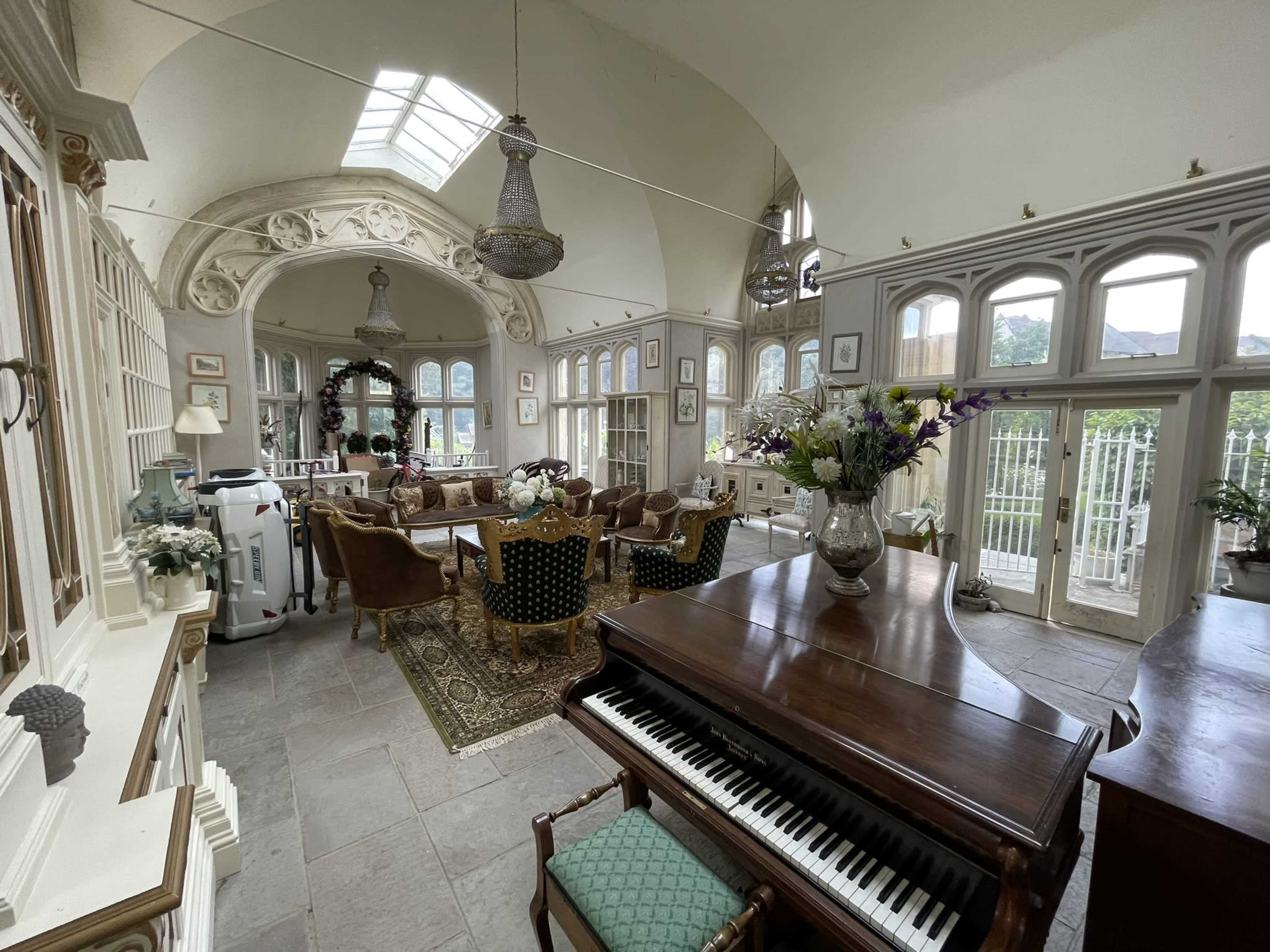 The image shows a spacious, elegantly designed room with large windows, intricate architectural details, a grand piano, and a mix of seating areas featuring decorative furniture and floral arrangements.
