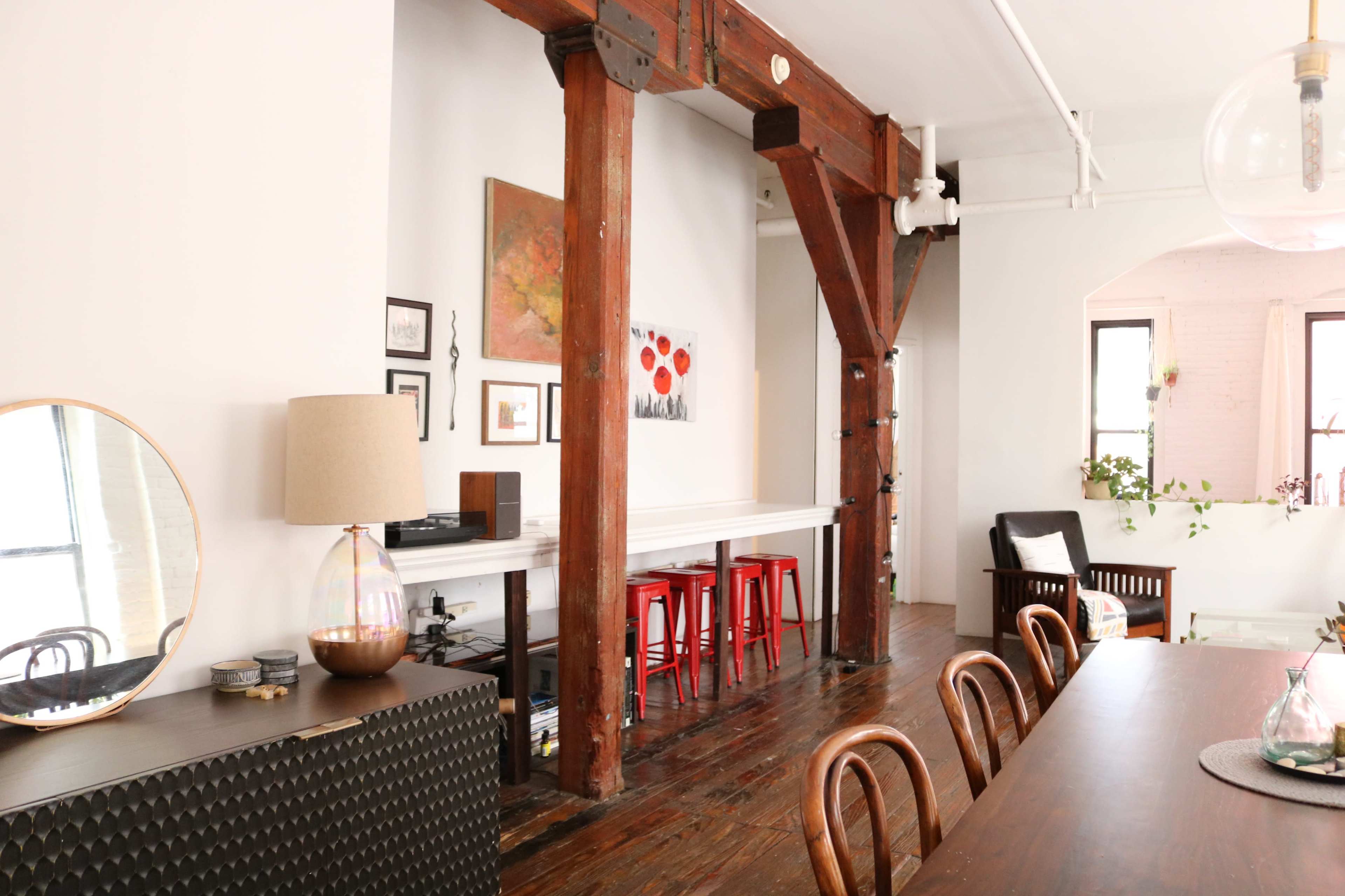An interior space featuring wooden beams, a dining area with a long table, and a small bar with red stools against white walls.