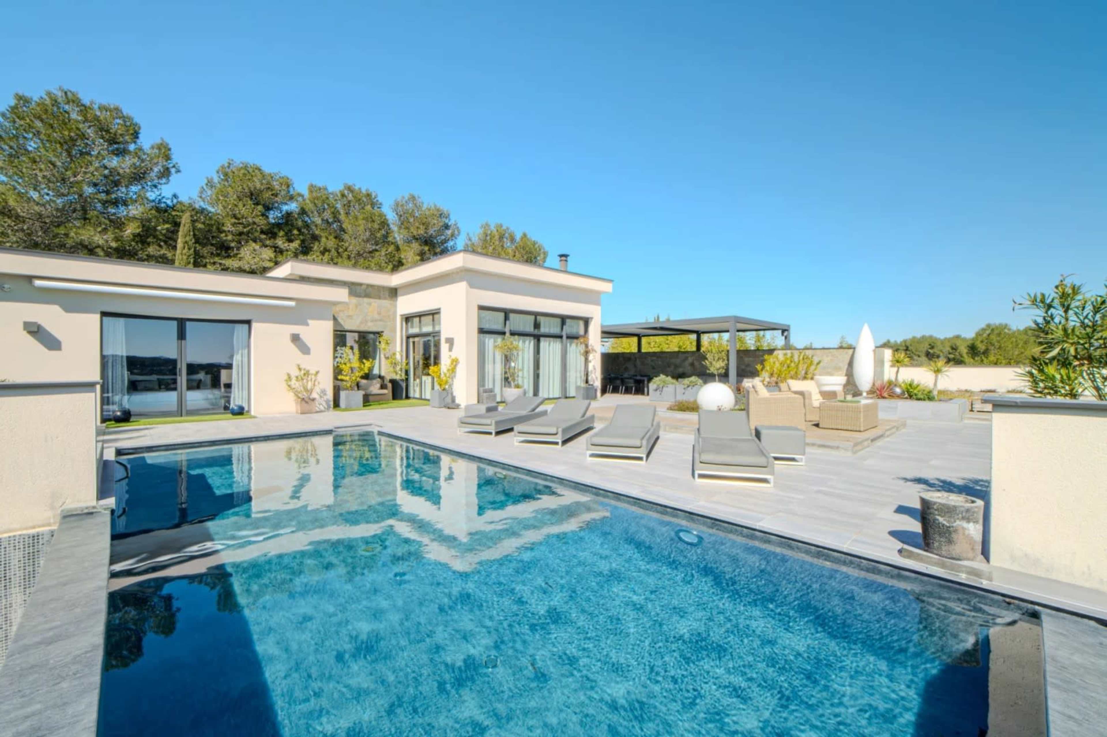 The image shows a modern home with a swimming pool surrounded by lounge chairs and lush greenery under a clear blue sky.