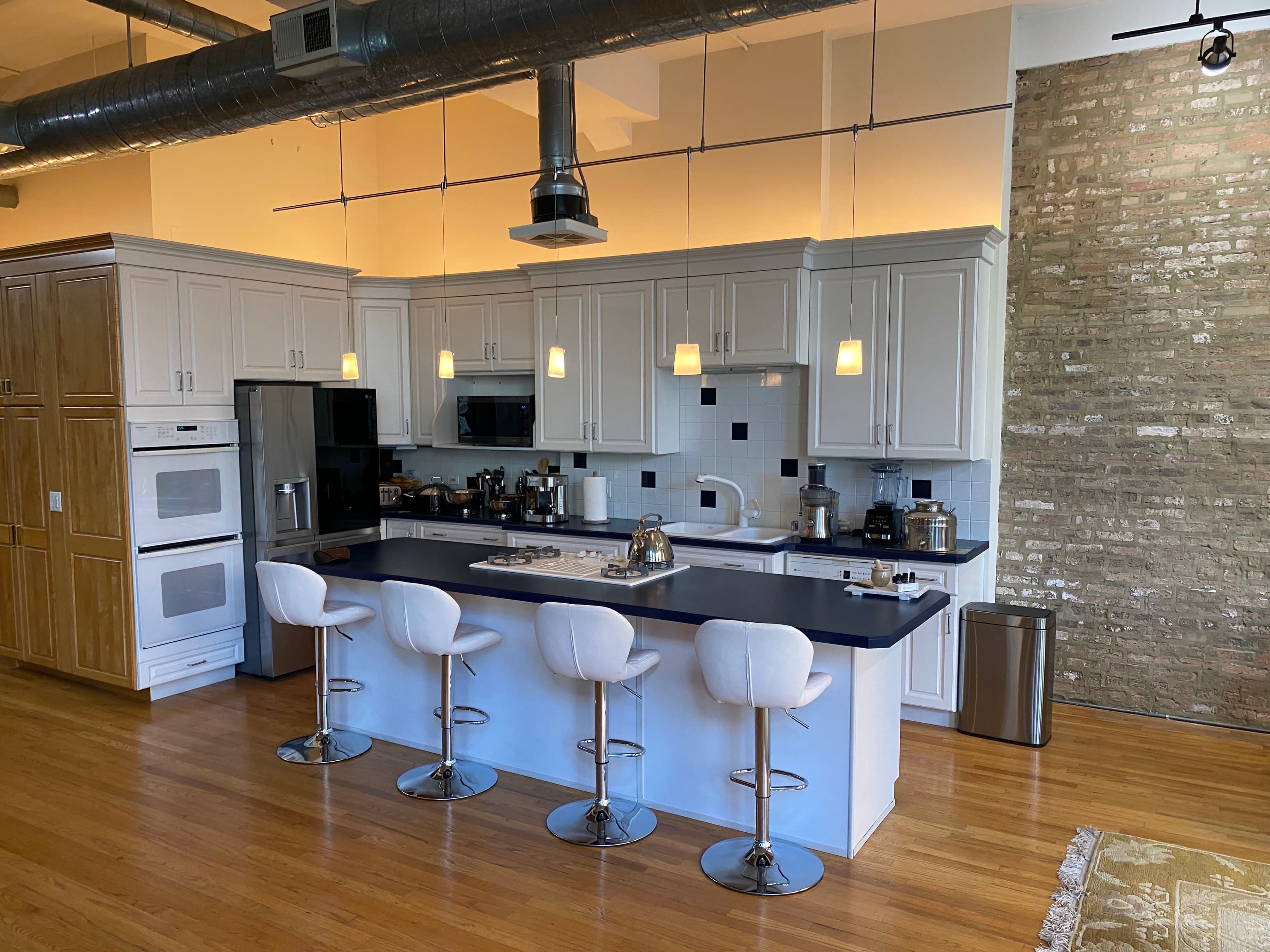A modern kitchen with a central island, four white bar stools, and stainless steel appliances, set against a brick wall.