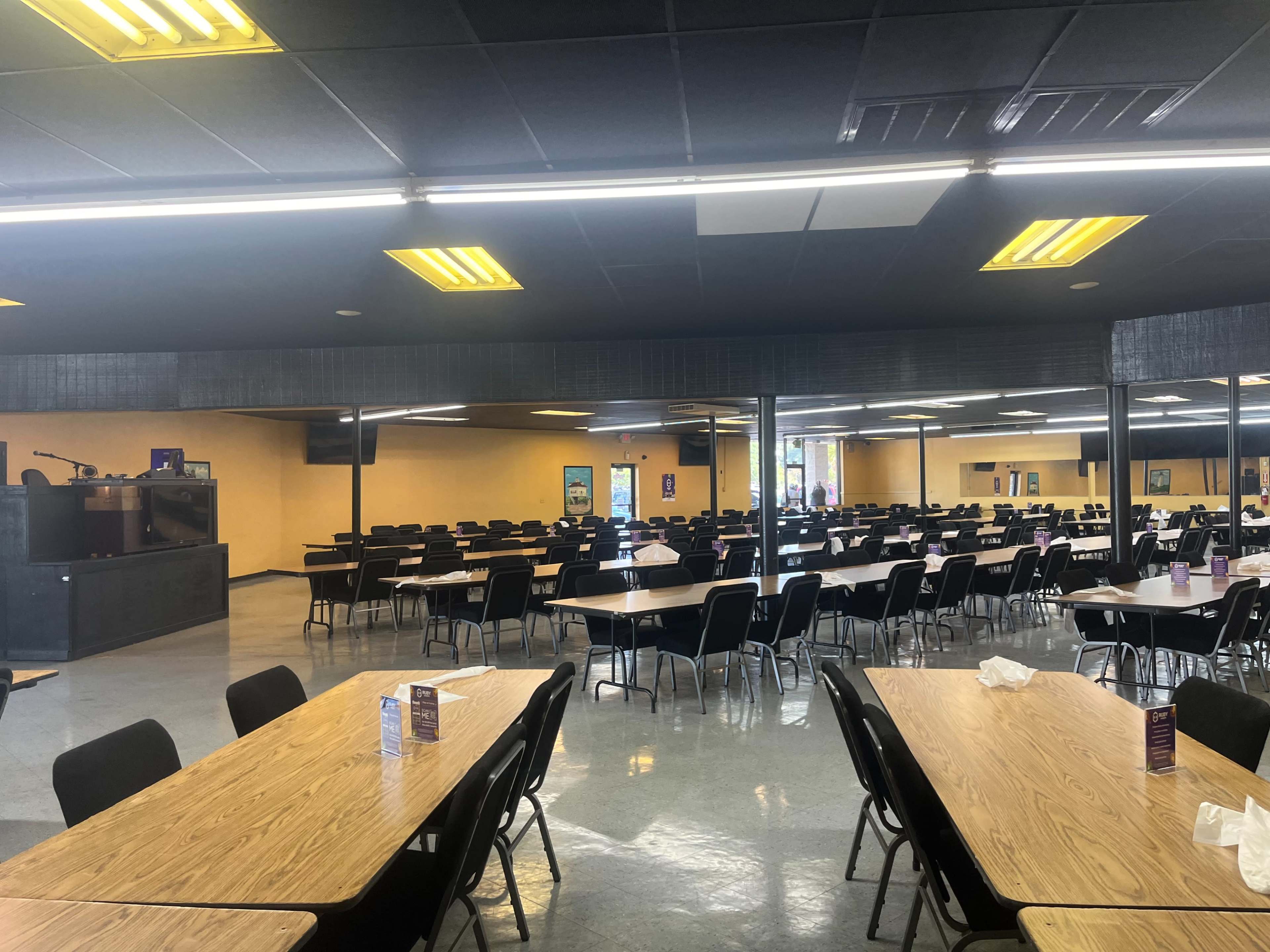 A large dining hall with multiple rows of wooden tables and black chairs arranged in a spacious, well-lit environment.