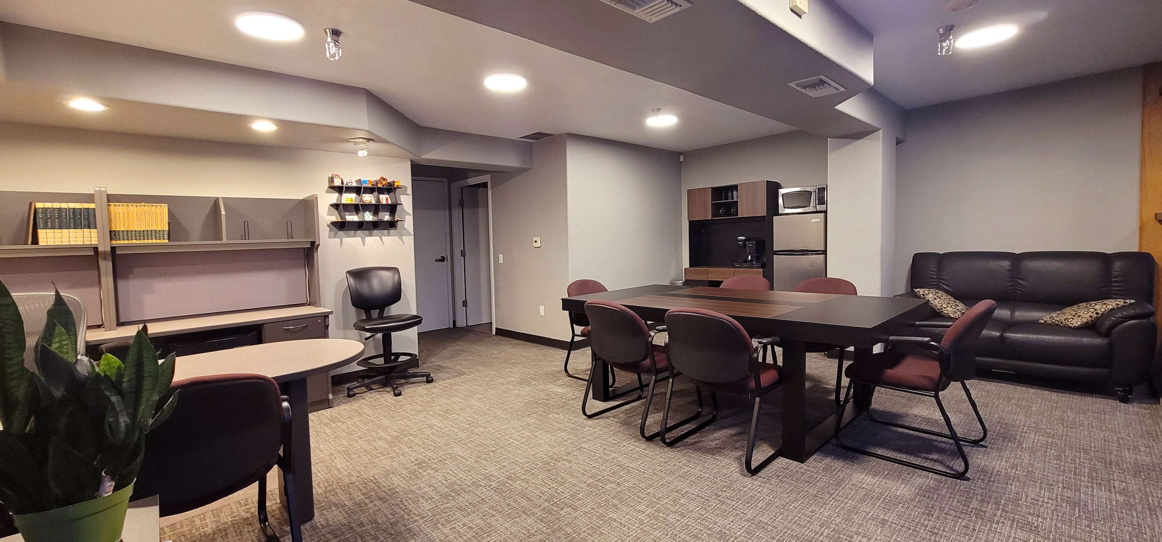 The image shows a modern office or meeting room with a large table, chairs, a couch, and kitchen appliances, all arranged in a well-lit space with neutral-colored walls.