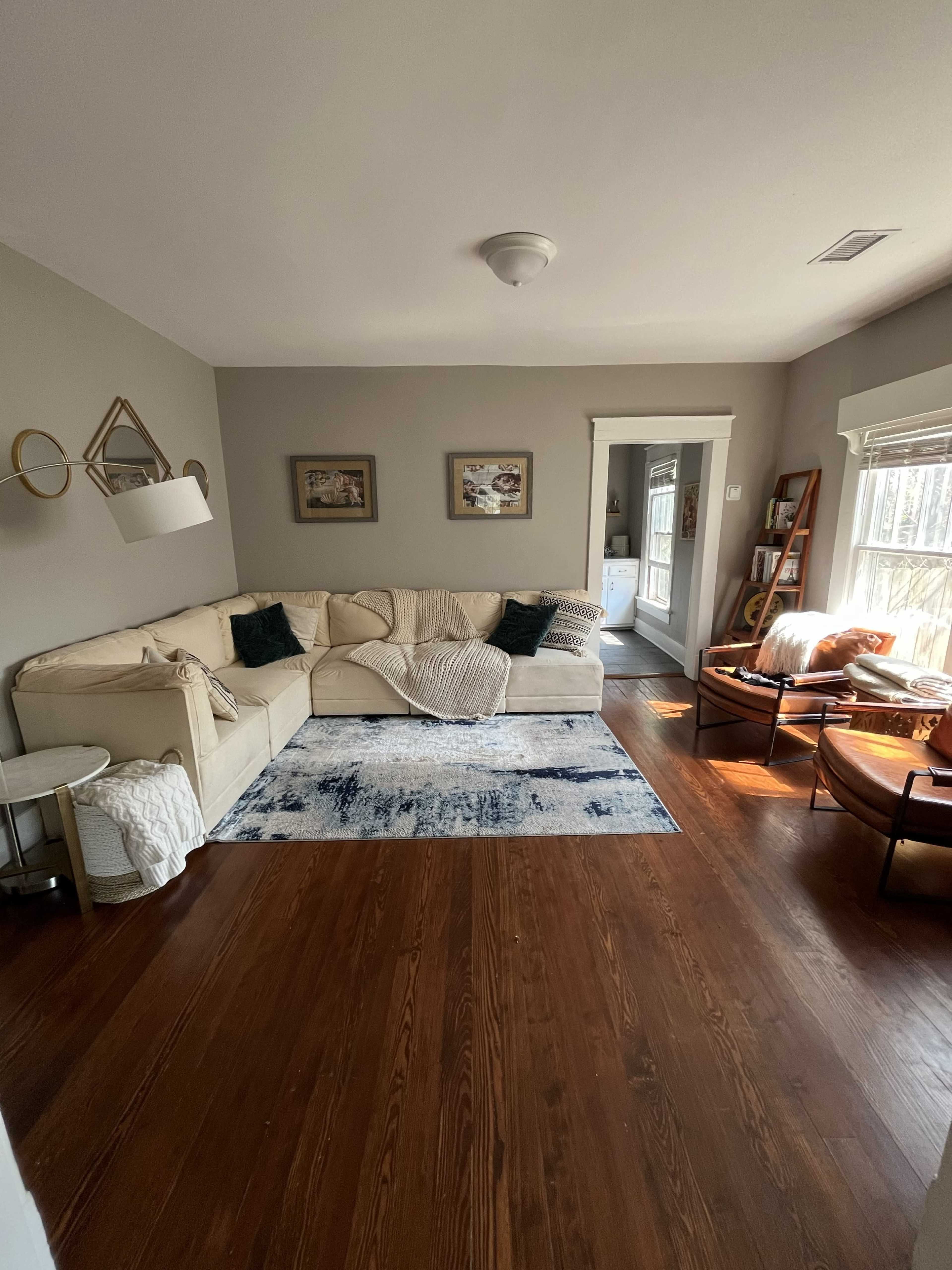 The image shows a living room with a light-colored sectional sofa, a decorative rug, wooden flooring, and a sunlit reading nook with two armchairs.