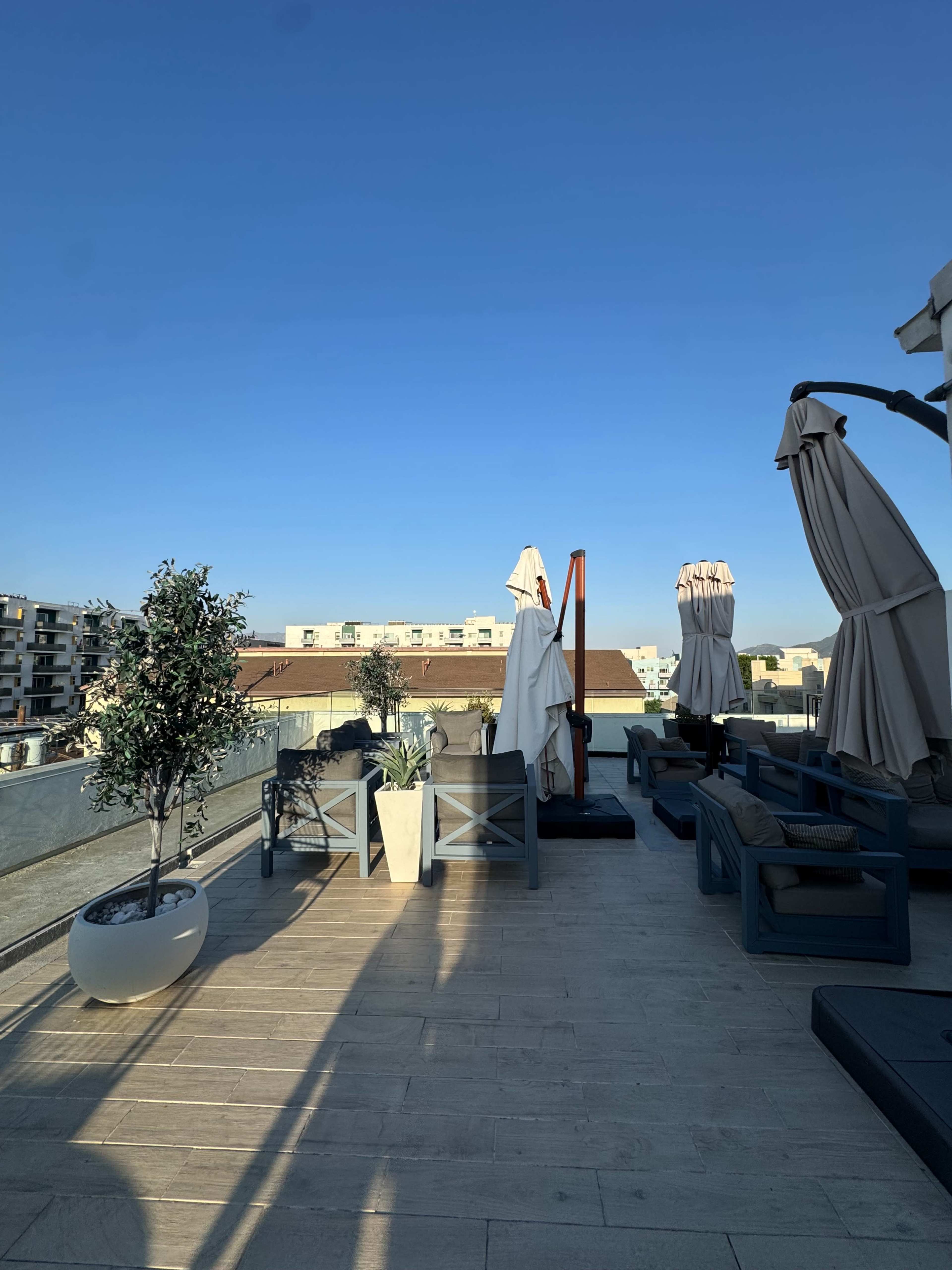 The image shows a rooftop terrace with lounge furniture, potted plants, and umbrellas under a clear blue sky.