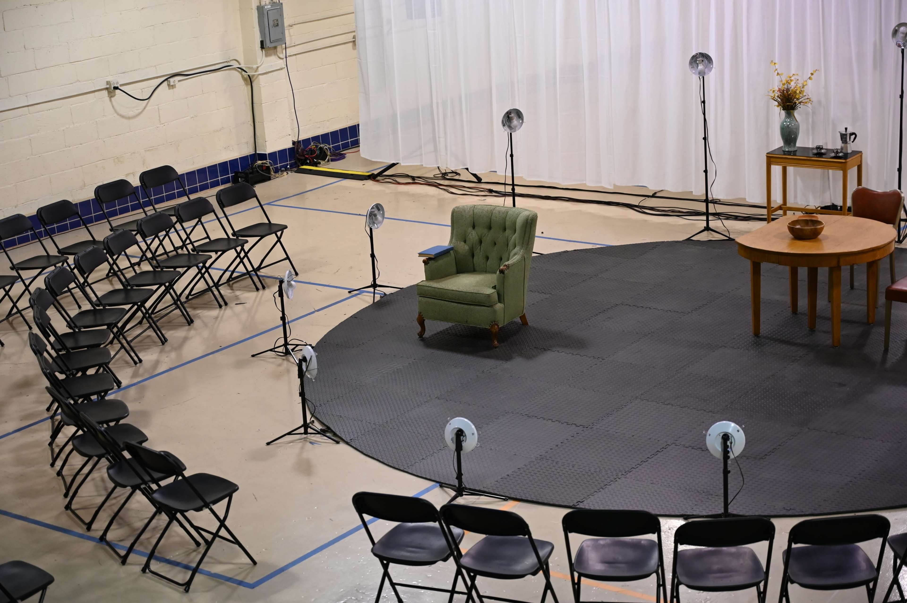 The scene shows a staged seating arrangement with black chairs facing a circular platform that features a green armchair and a wooden table with a vase.