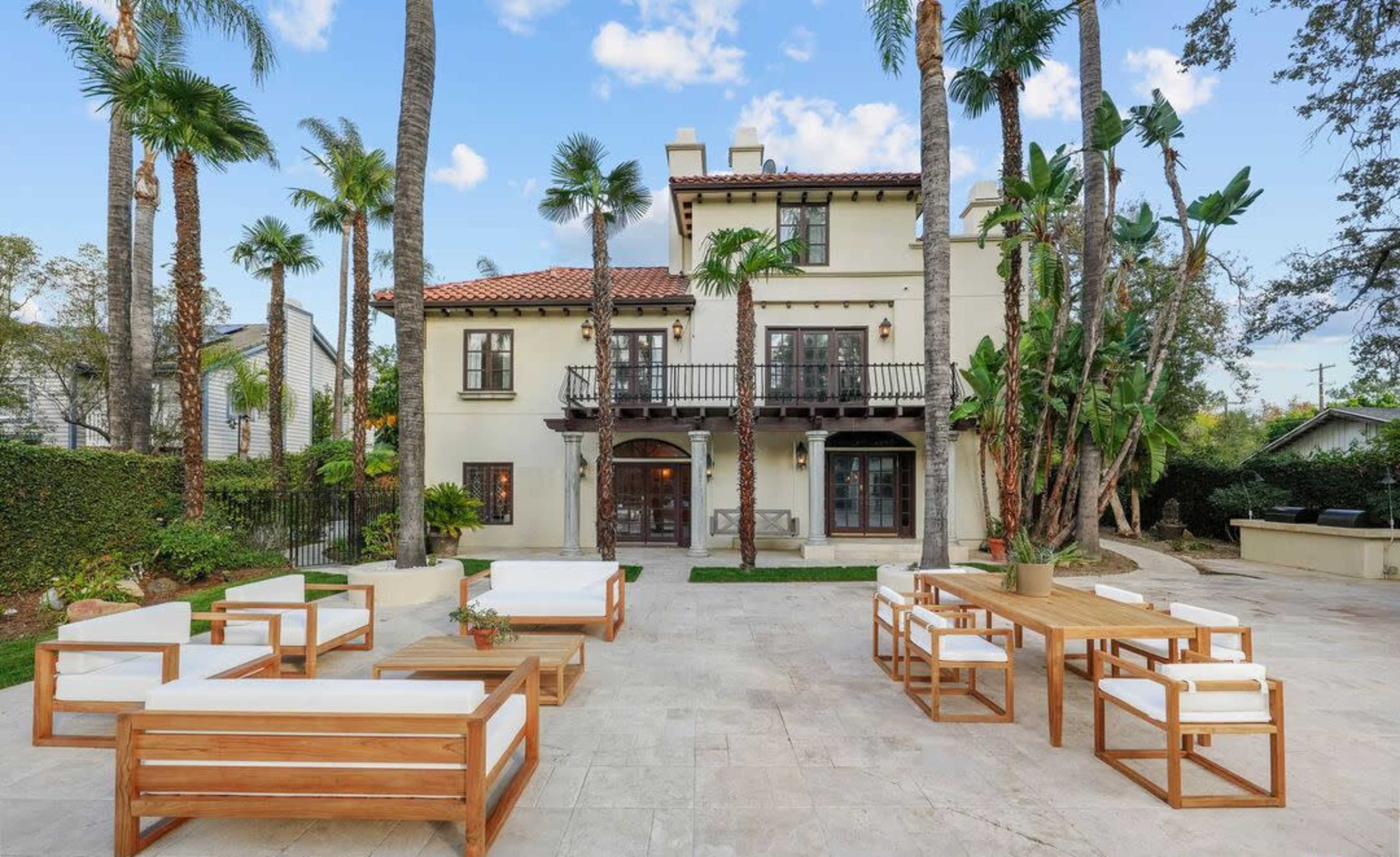 The image shows a modern house with a tile roof surrounded by palm trees, featuring a spacious patio area furnished with wooden seating and a dining table.