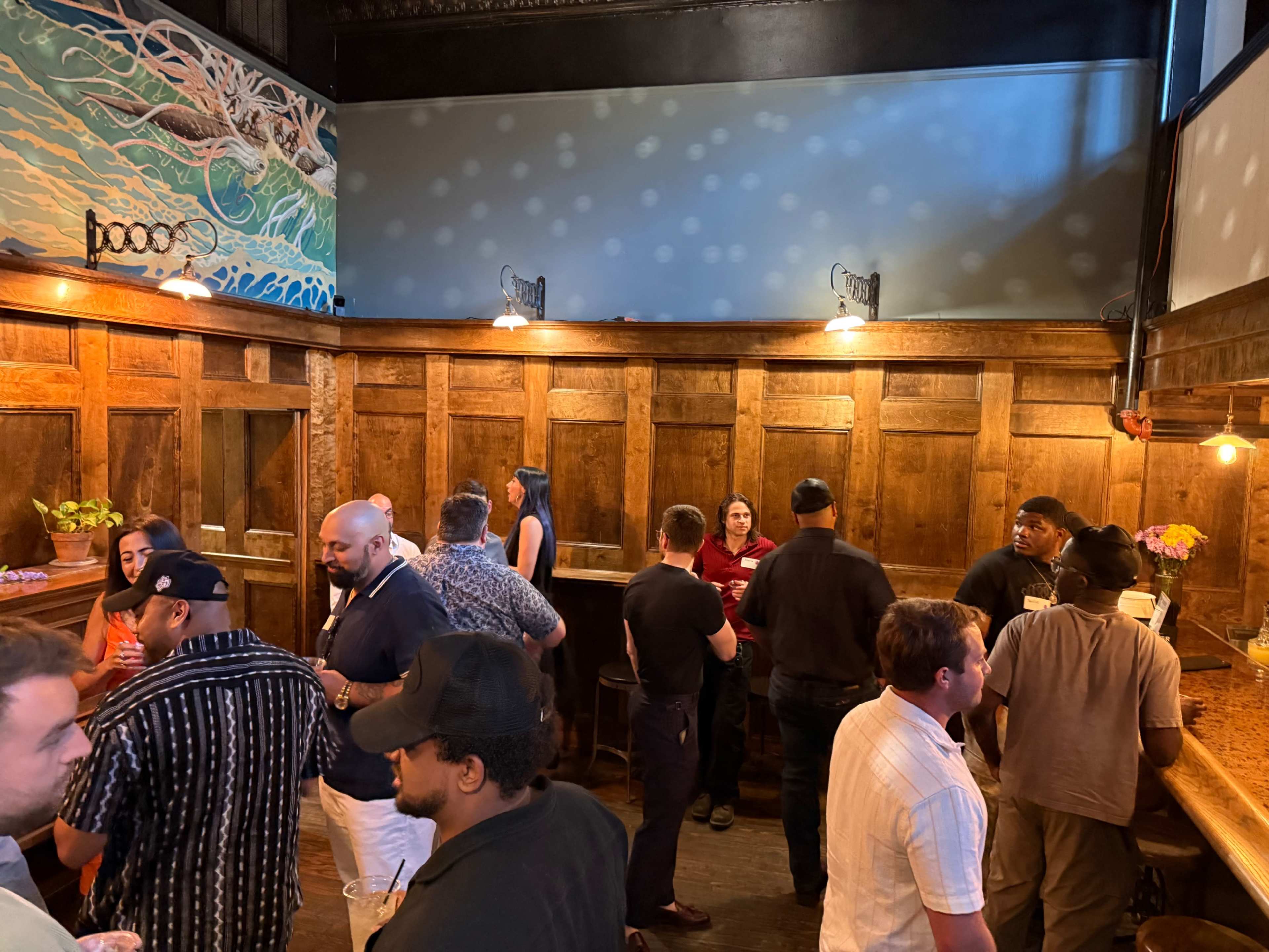 A group of people socialize in a dimly lit bar with wooden walls and a decorative mural on one side.