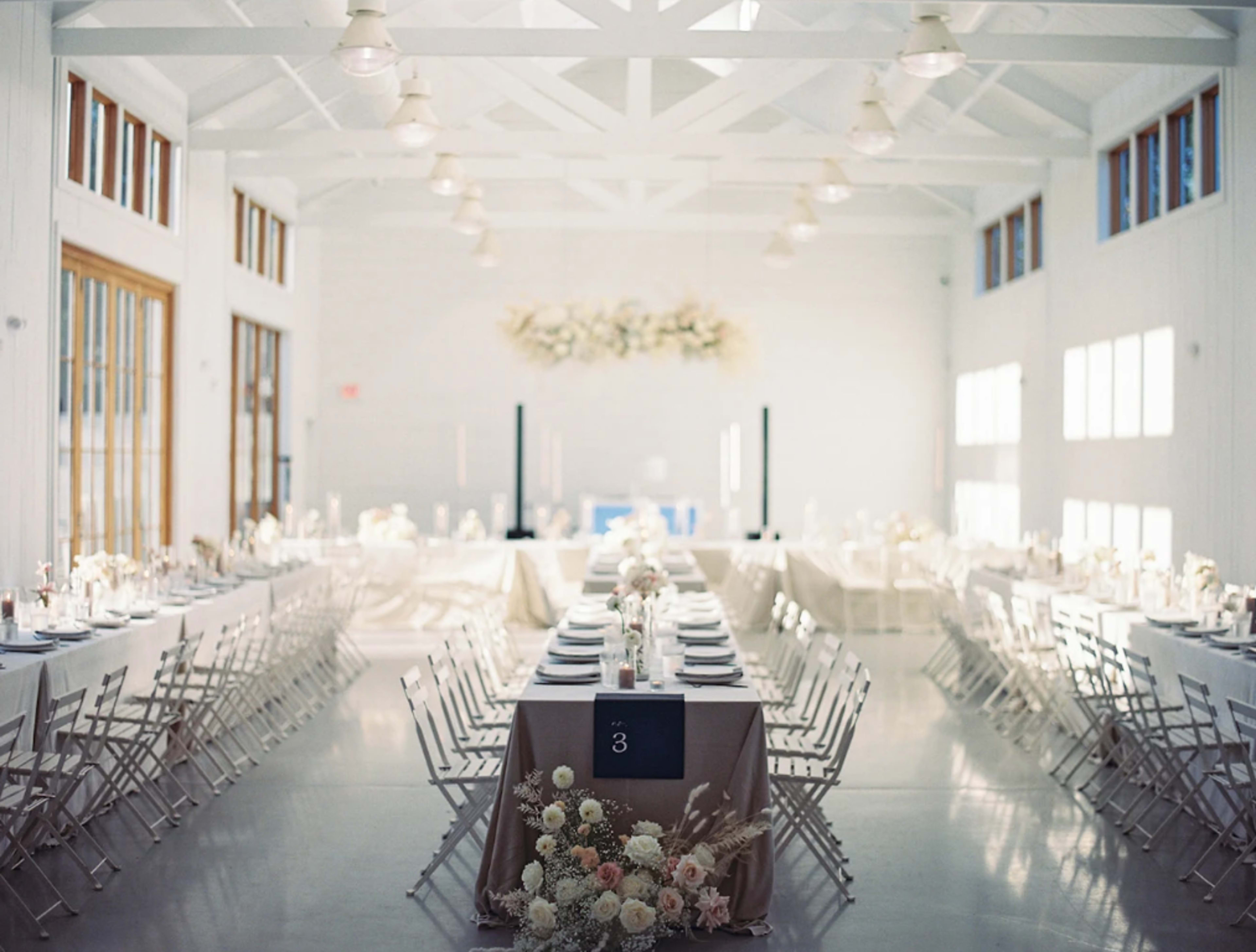 A spacious, elegantly decorated dining hall features long tables set for a formal event, with flowers as centerpieces and white walls enhancing the bright atmosphere.