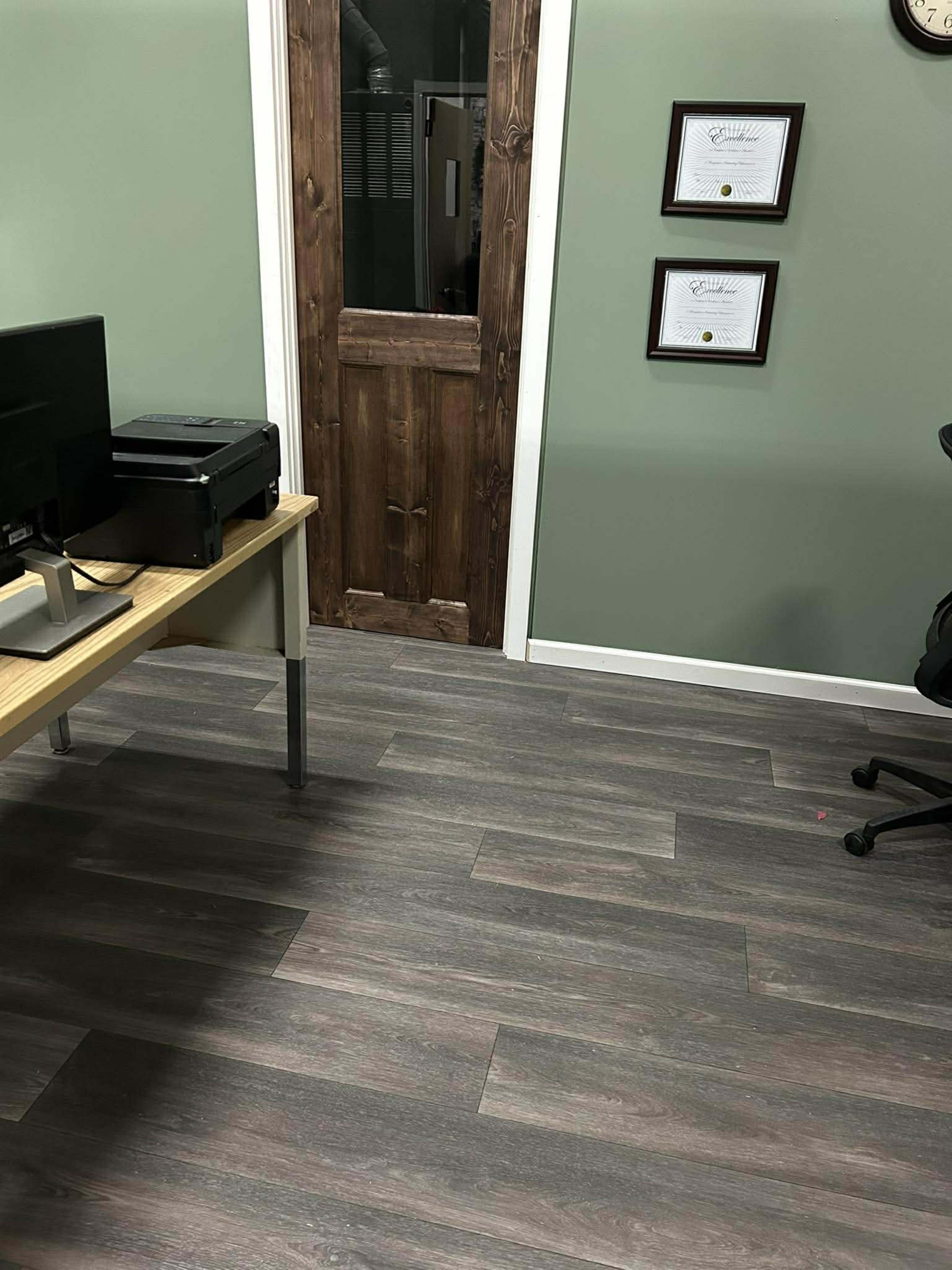 The image shows an office space with a wooden door, a printer on a desk, and framed certificates on the wall.