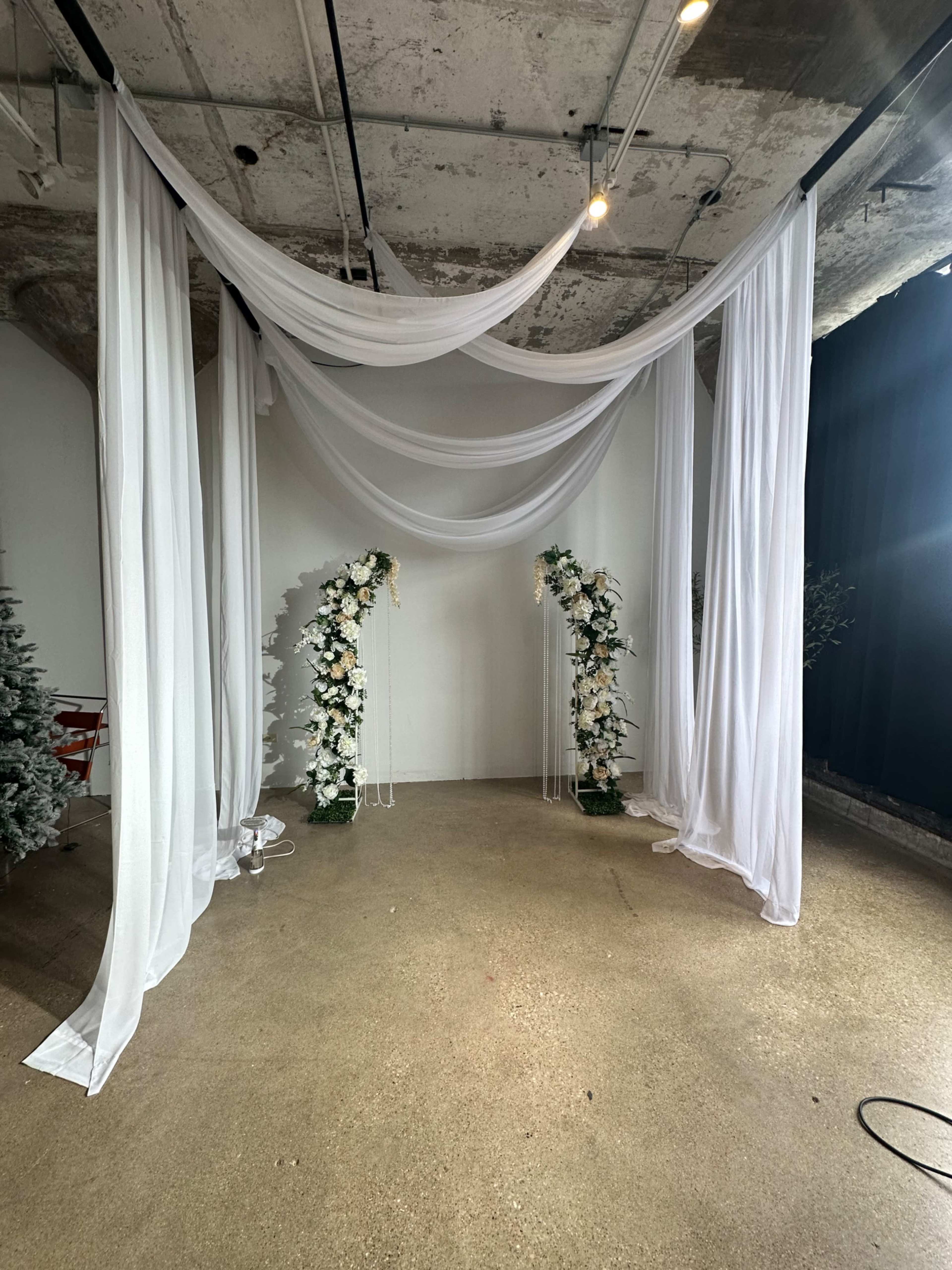 The image shows a decorated indoor space with white draped fabric and floral arrangements on either side, creating a backdrop that looks suitable for a wedding or event ceremony.