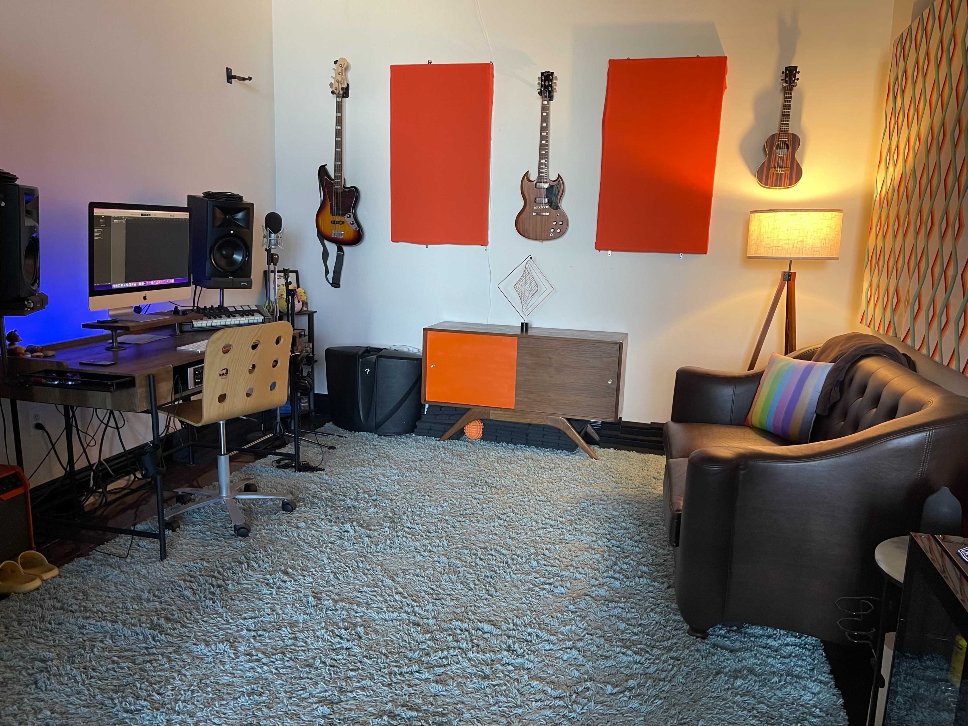 A music studio features a desk with a computer and keyboard, two guitars hanging on the wall, a brown couch, and a colorful patterned artwork above a side table.