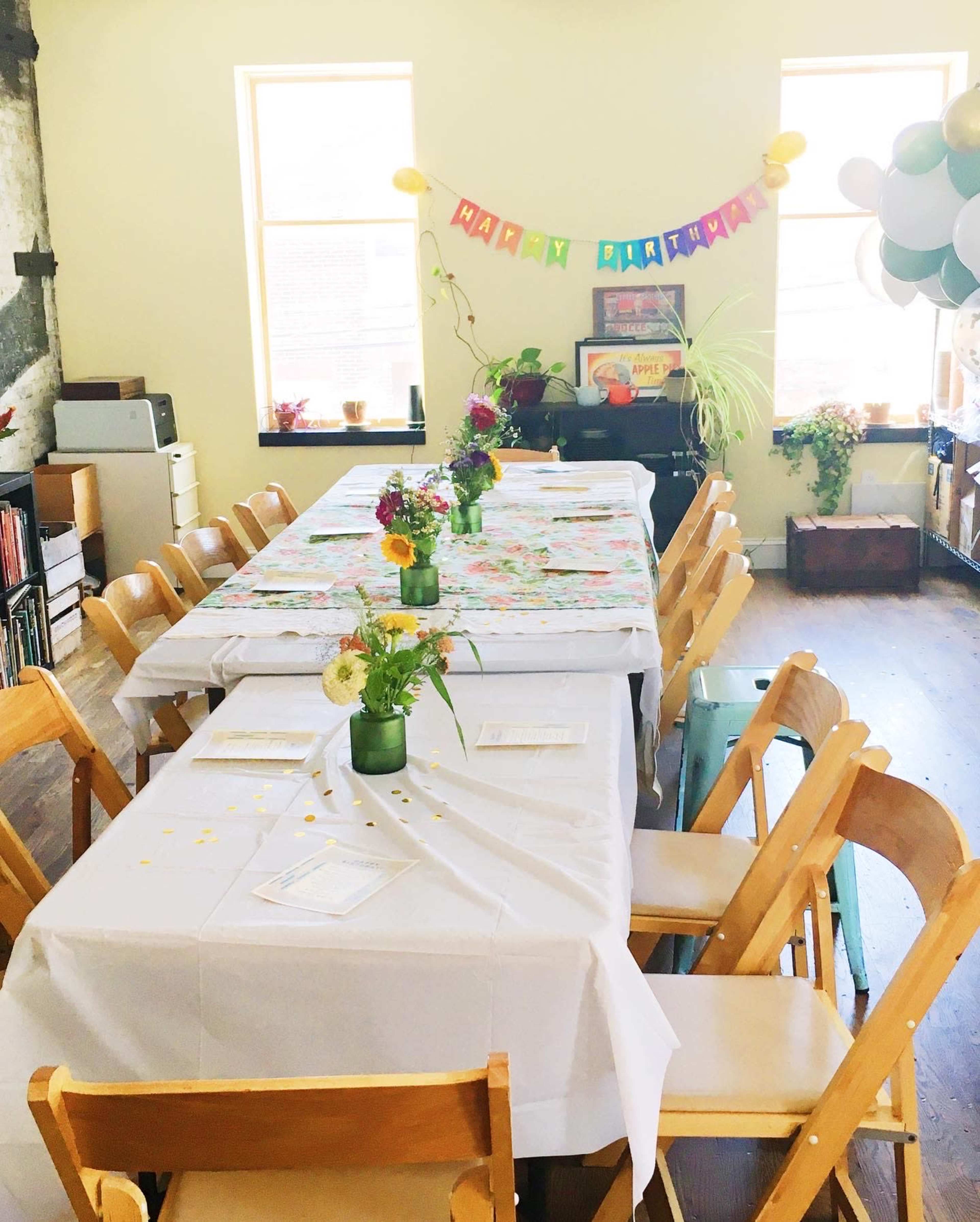 A decorated indoor space with two long tables covered in white tablecloths and surrounded by wooden chairs, featuring colorful birthday decorations and flower centerpieces.
