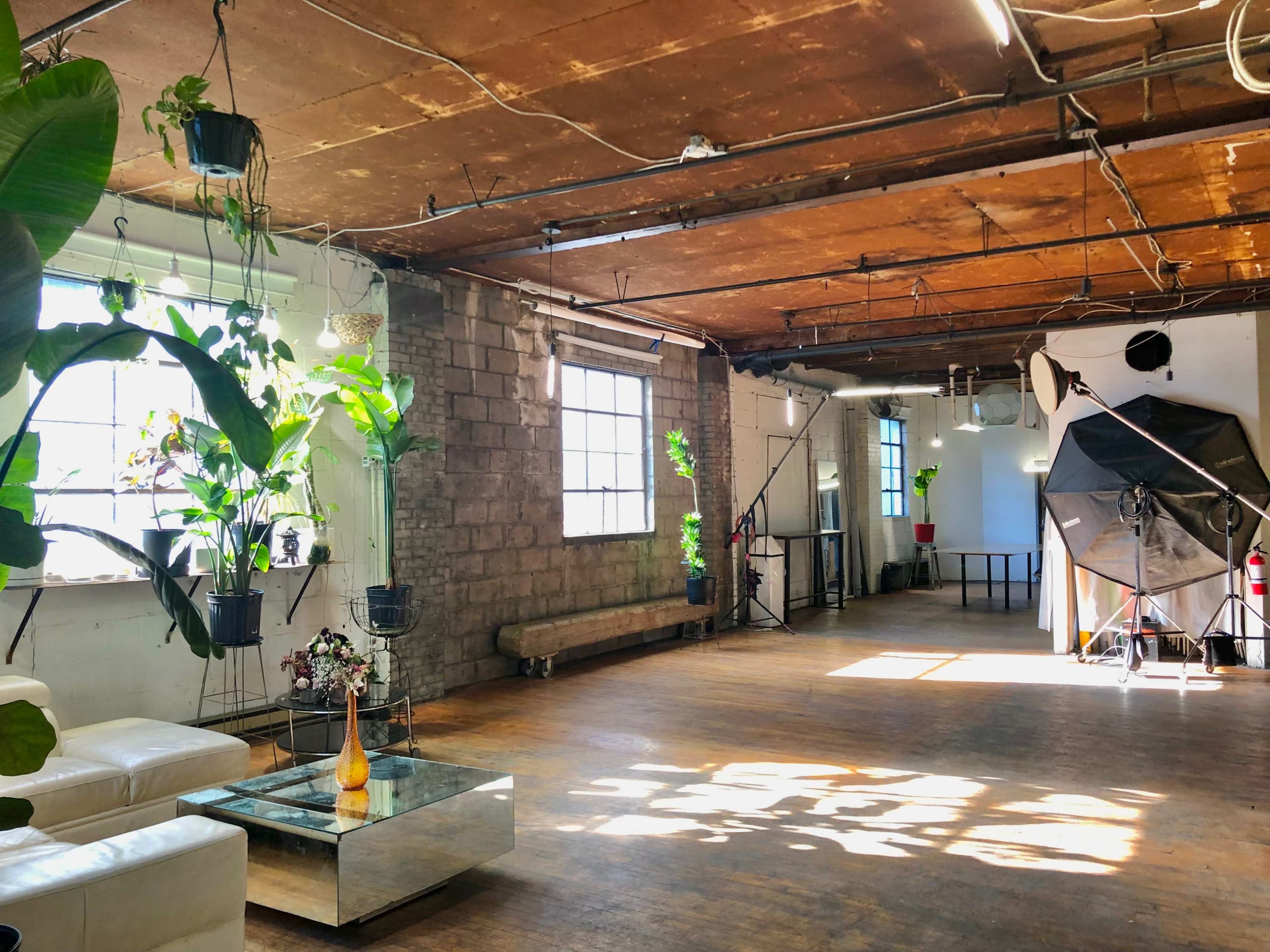 The image shows a spacious, industrial-style room with exposed brick walls, large windows, and plants hanging from the ceiling, along with a white couch and a glass coffee table in the center.
