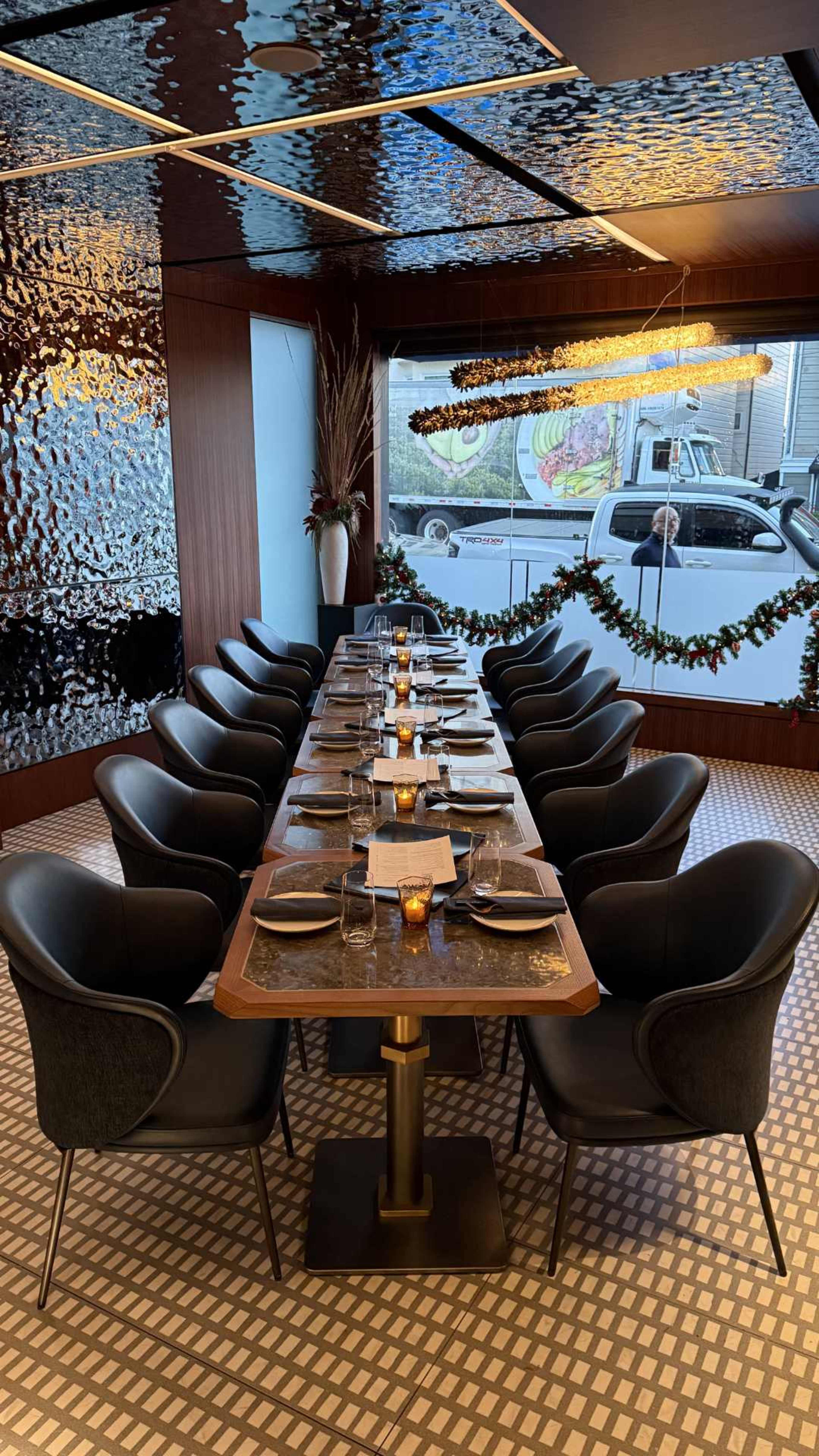 A long dining table with black chairs is set for a meal in a modern restaurant, featuring a decorative ceiling and window view of an outside street.