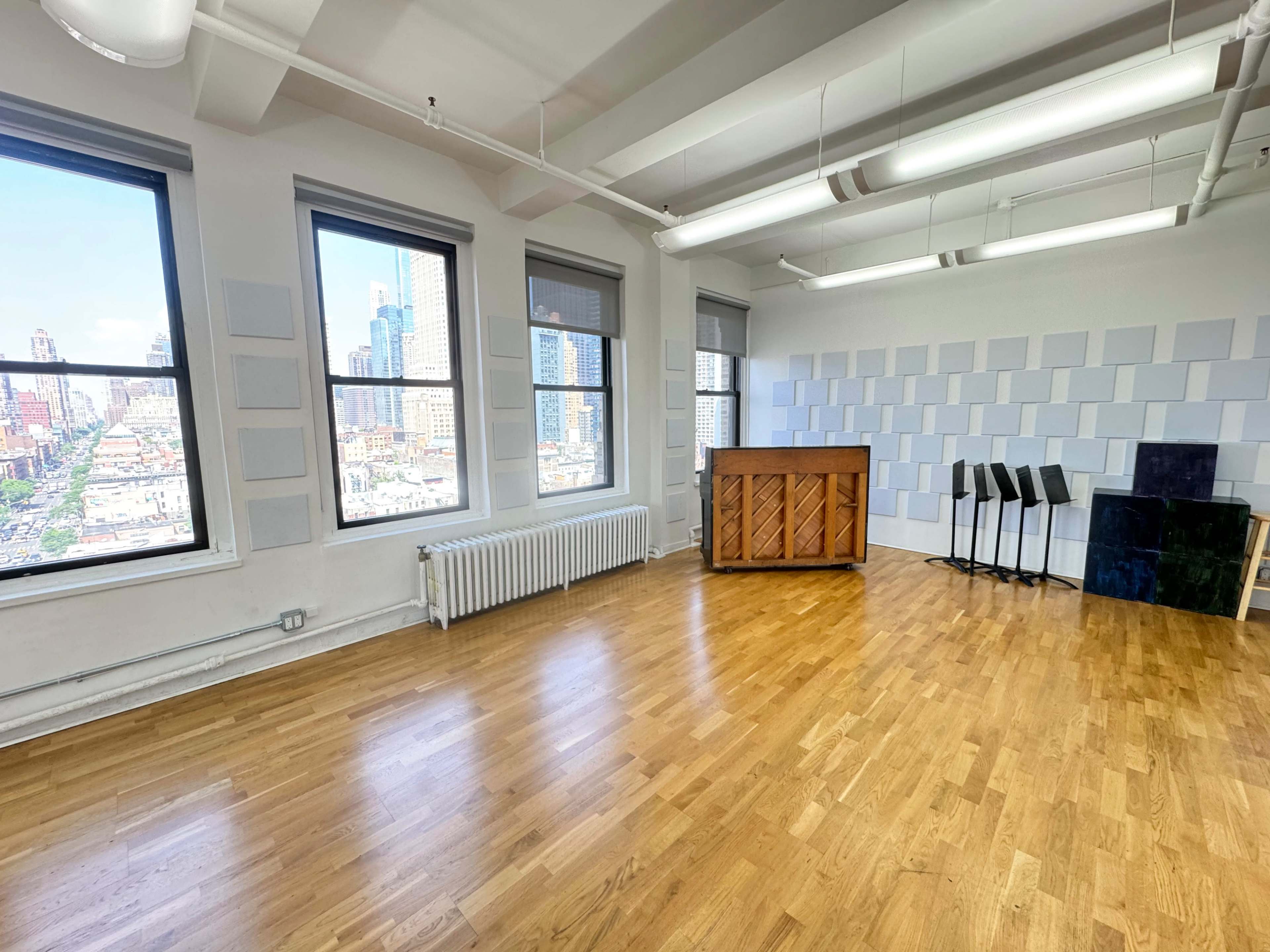 A spacious, well-lit room with large windows, a wooden piano, and several music stands against a white wall.