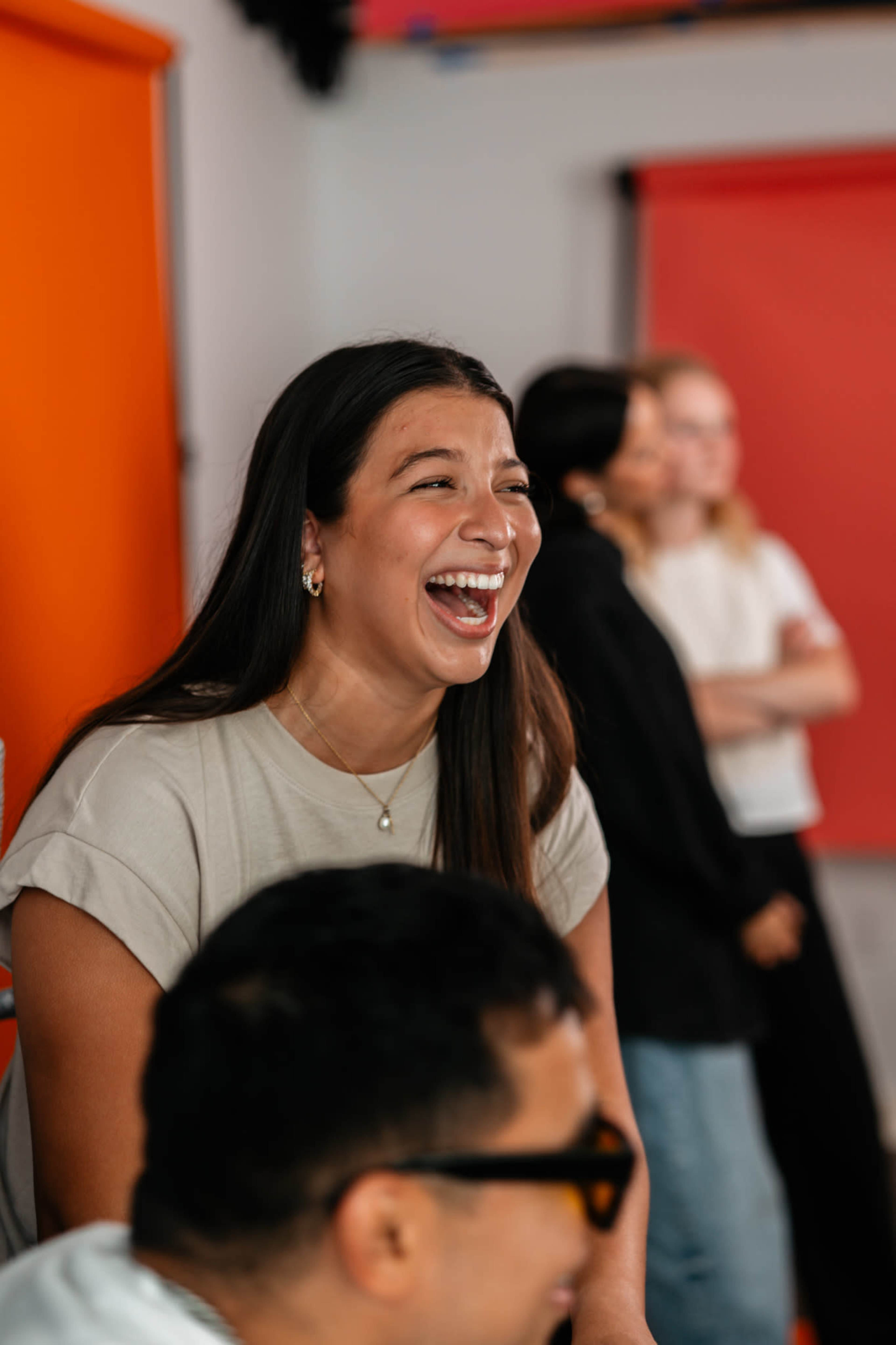 A woman with long hair laughs while sitting in a room with orange backgrounds, as others stand in the background.