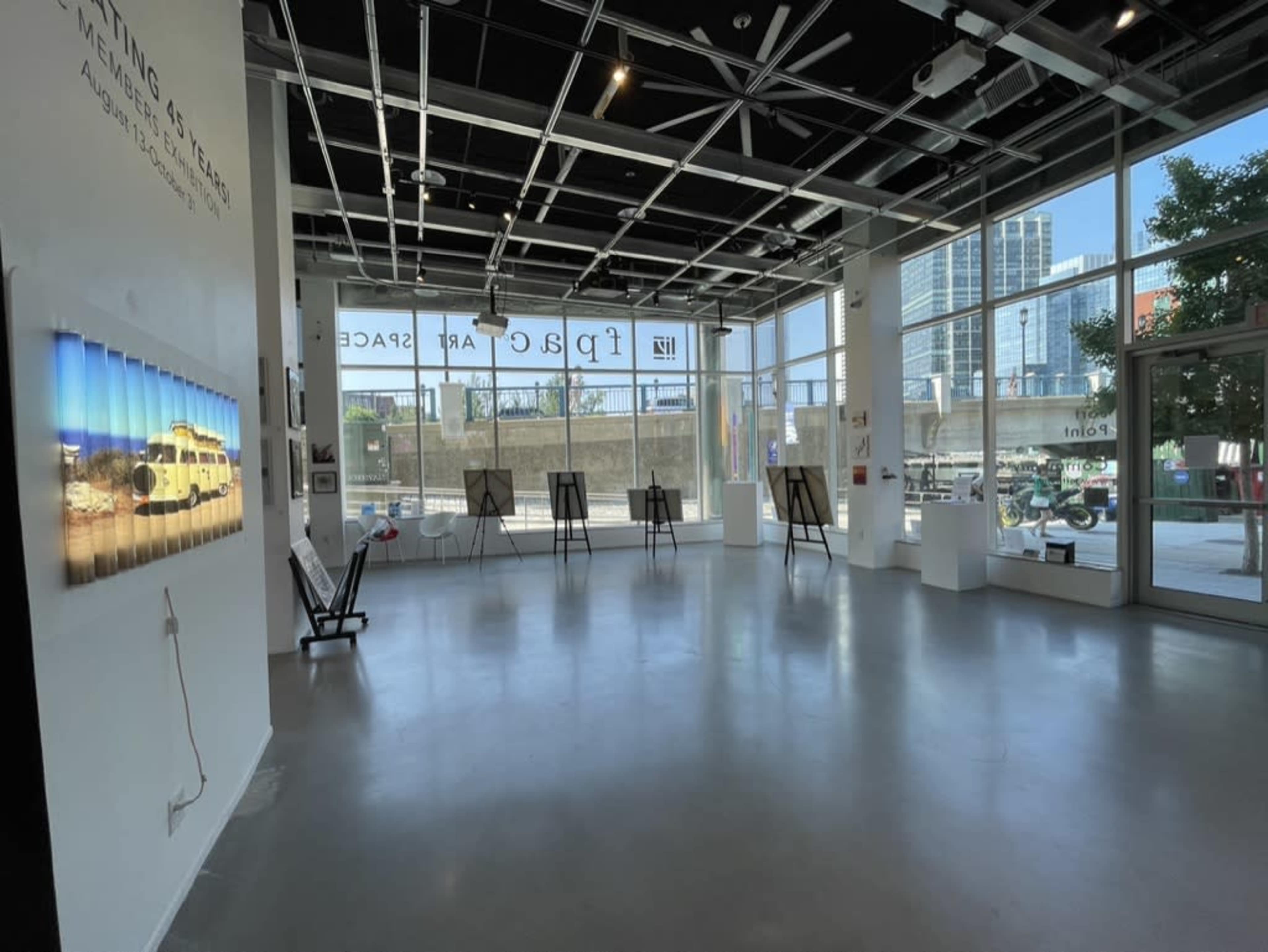 A spacious art gallery features large windows, empty easels, and a photograph on the wall, with cityscape visible outside.