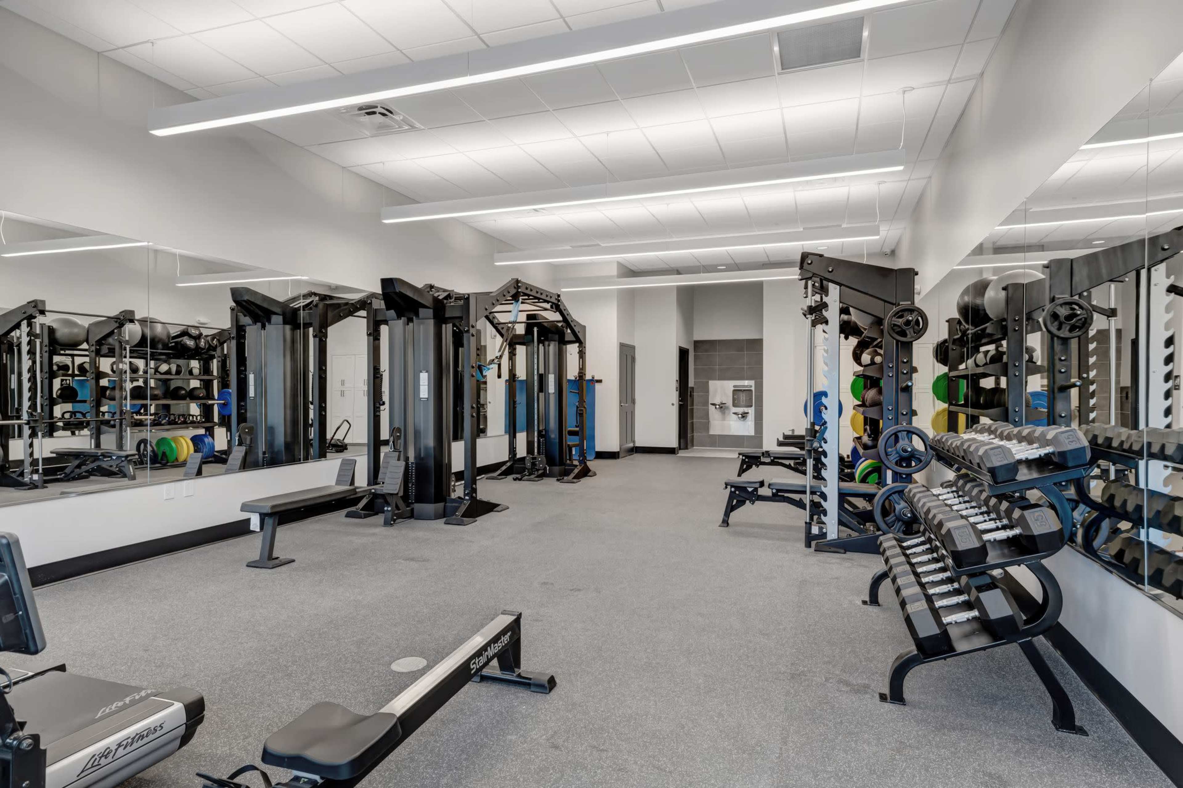 The image shows a well-equipped gym with weight machines, free weights, and a treadmill arranged in a spacious, brightly lit area.