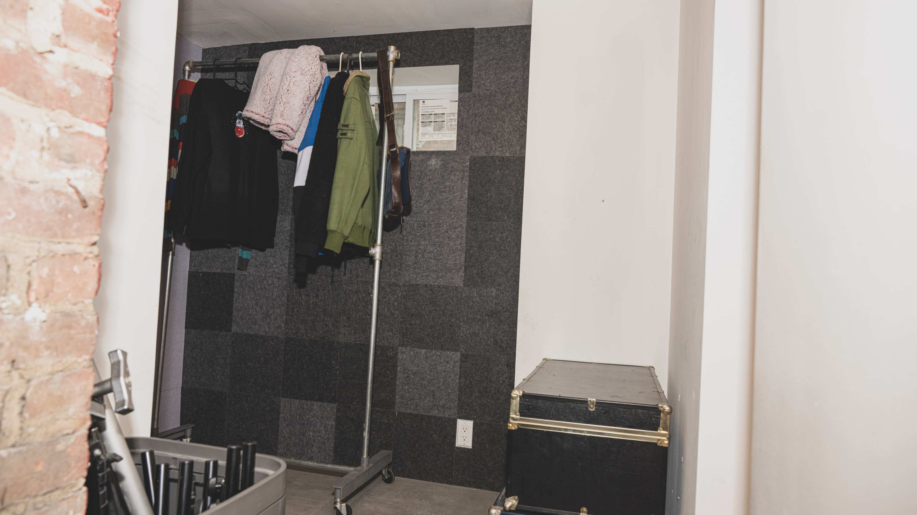 The image shows a small closet space with a clothing rack displaying several garments against a gray, patchwork wall, alongside a storage trunk in the corner.