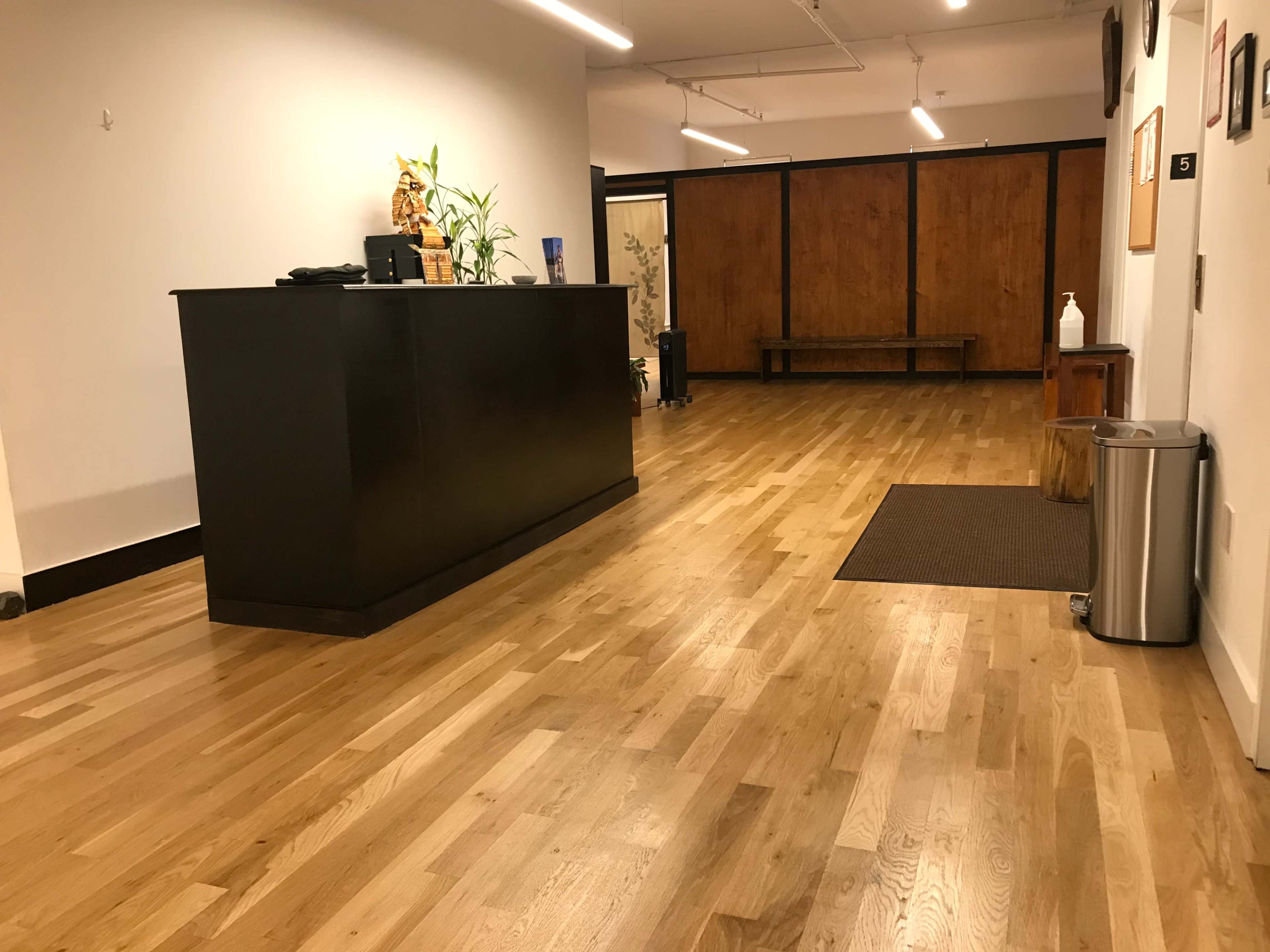 The image shows a spacious lobby area with wooden flooring, a reception desk on the left, and a brown wall in the background.