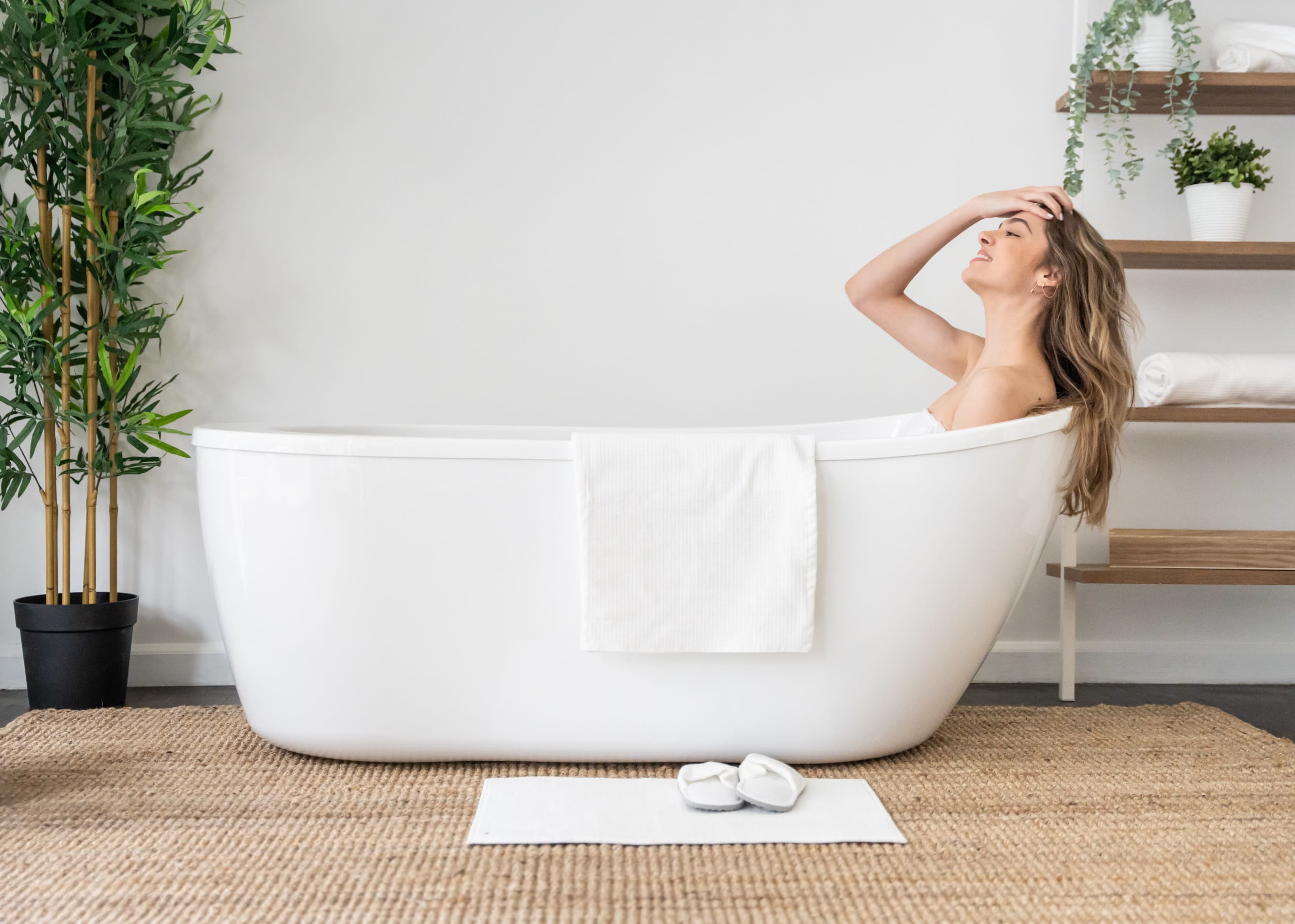 A woman relaxes in a white bathtub with a towel draped over the side, surrounded by greenery and a wooden shelf.