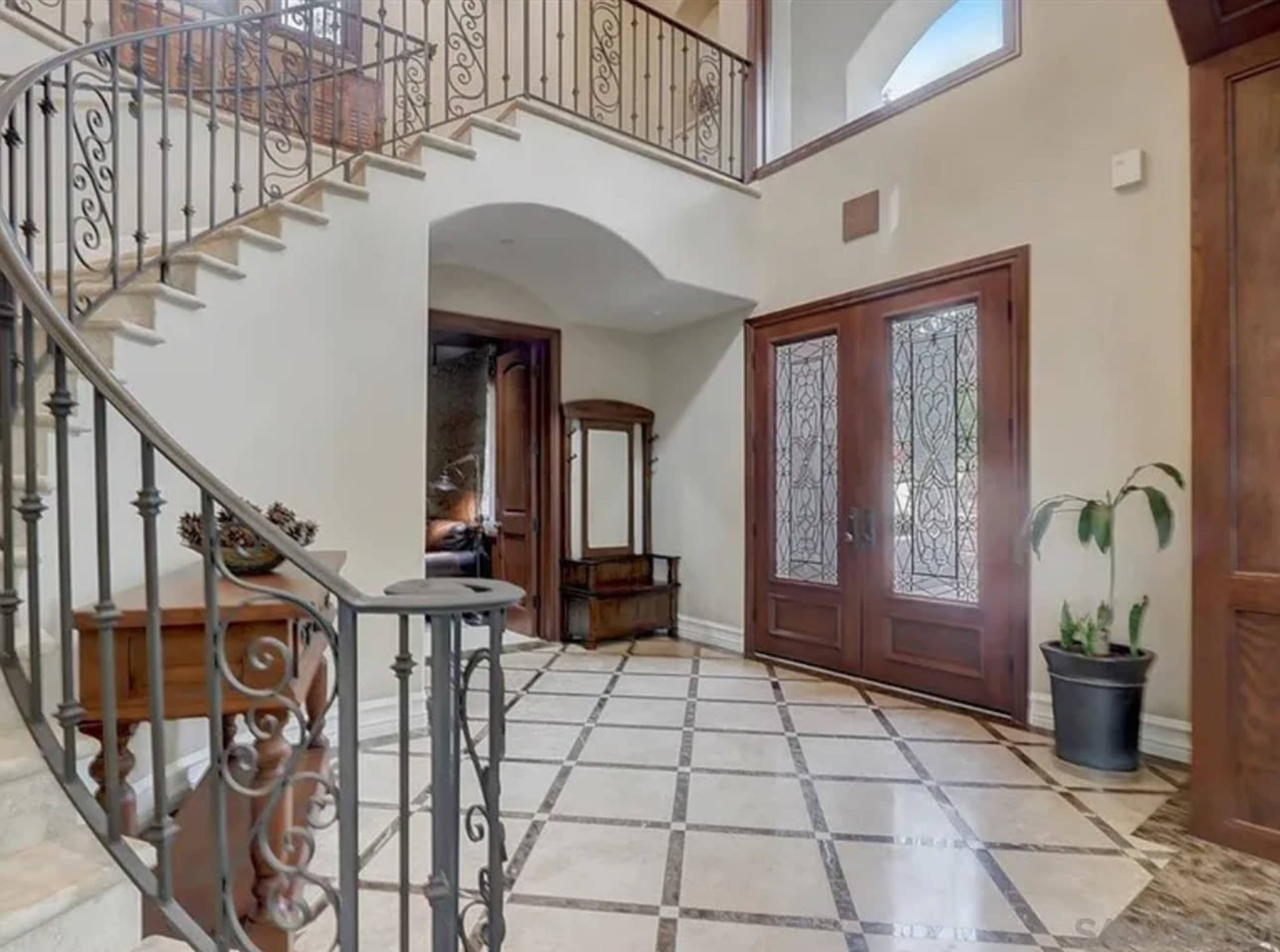 The image shows a foyer with a curved staircase, decorative iron railing, and double doors with glass panels opening into the space.