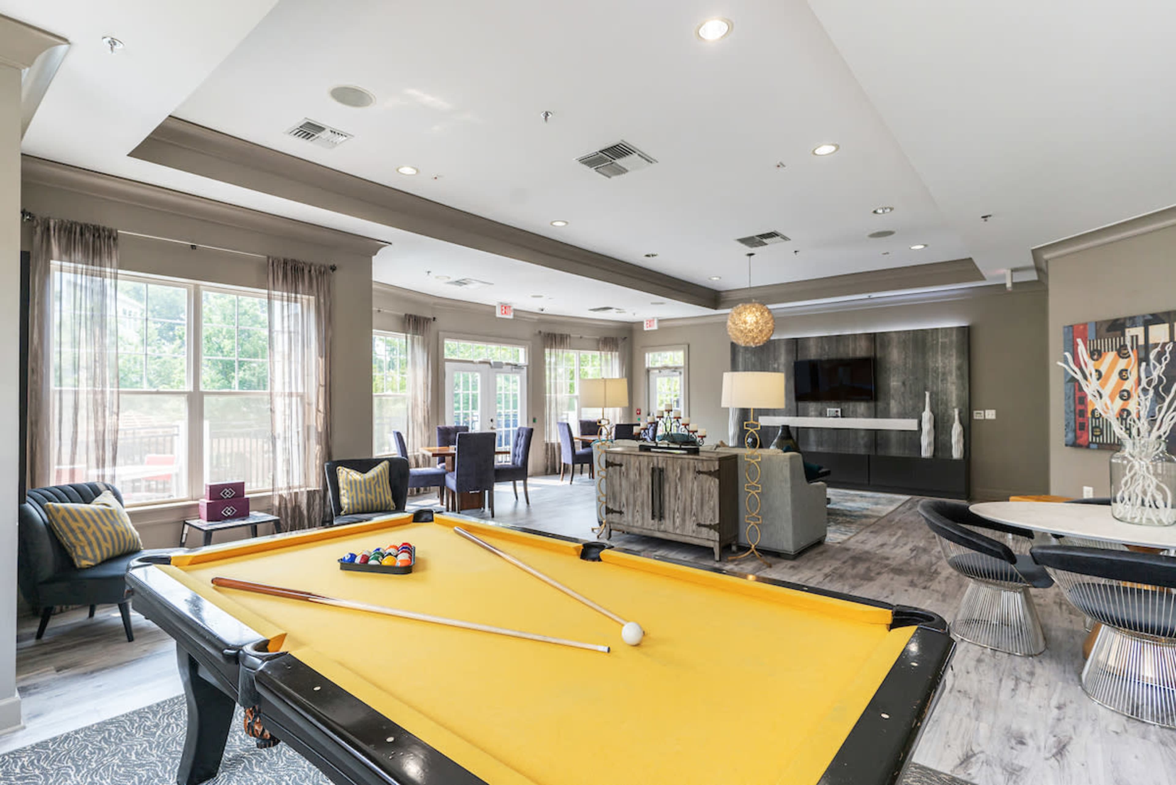 A brightly lit room features a pool table in the foreground, with seating areas and large windows in the background.
