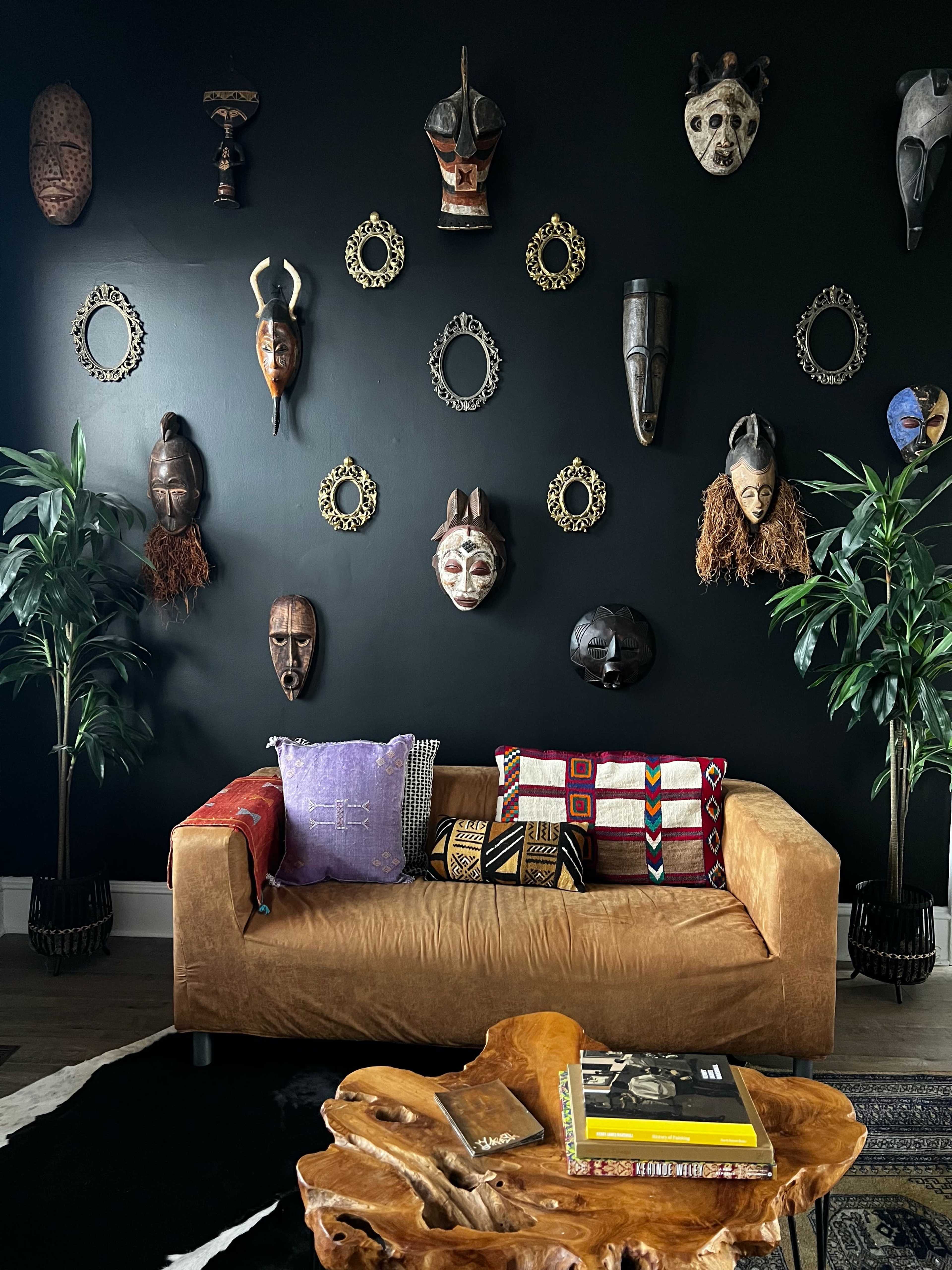 A living room features a brown sofa adorned with patterned cushions, a round wooden coffee table, and a wall decorated with various masks and mirrors against a black background.