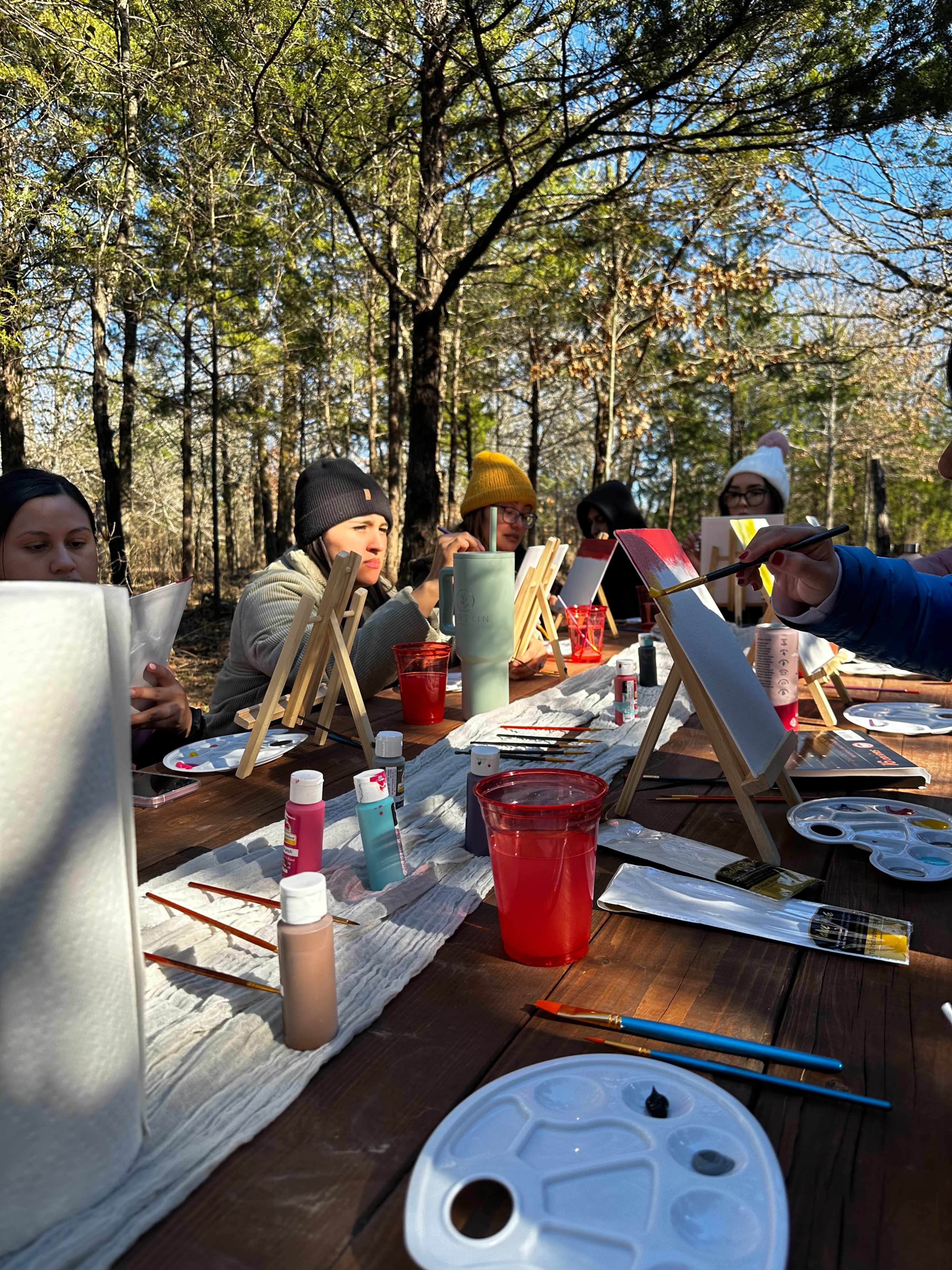 A group of people sits at a long wooden table in a forested area, engaged in a painting activity with easels, paint palettes, and cups of paint.