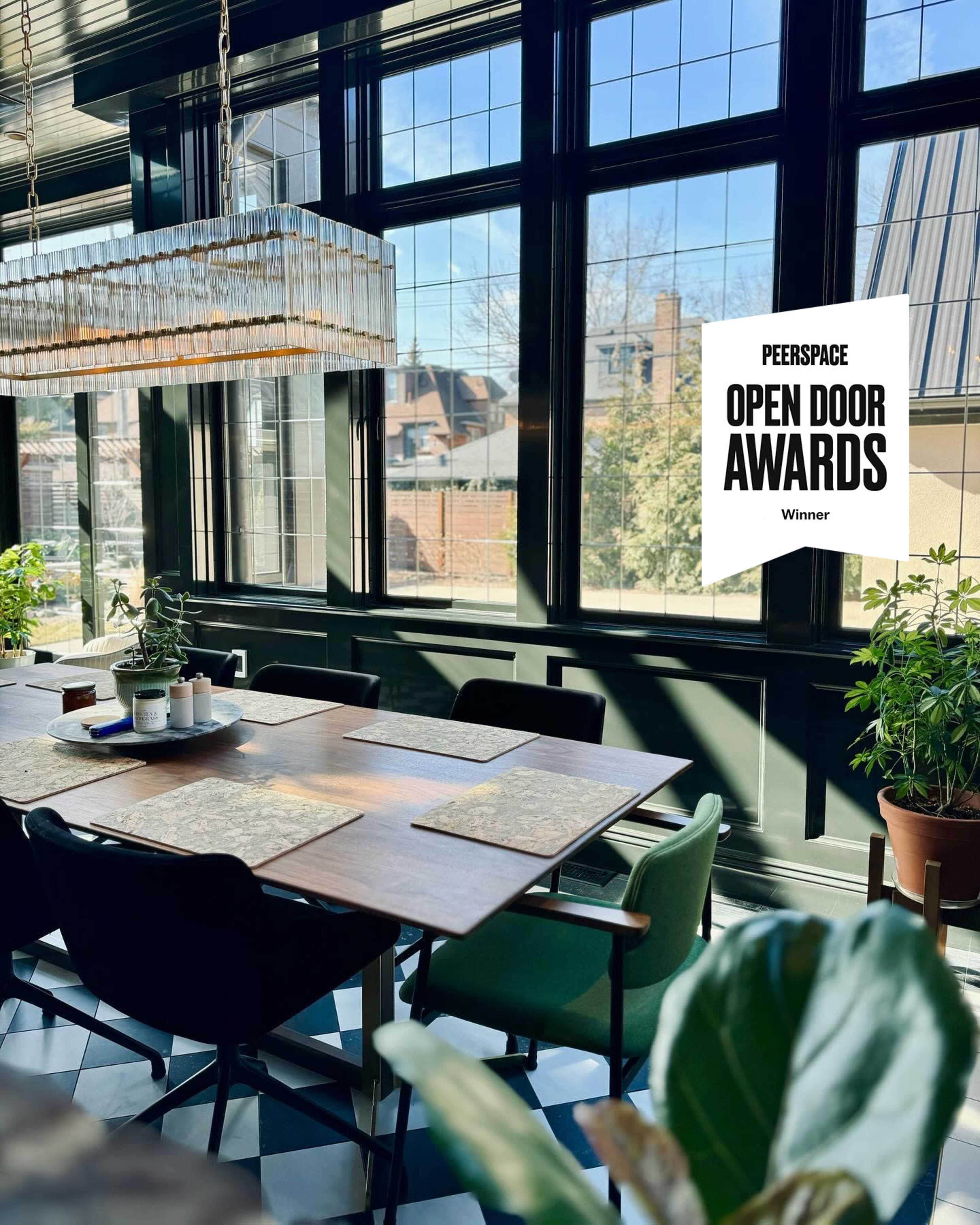 The image shows a stylish dining area with a large wooden table, surrounded by green chairs, large windows allowing natural light, and several indoor plants.