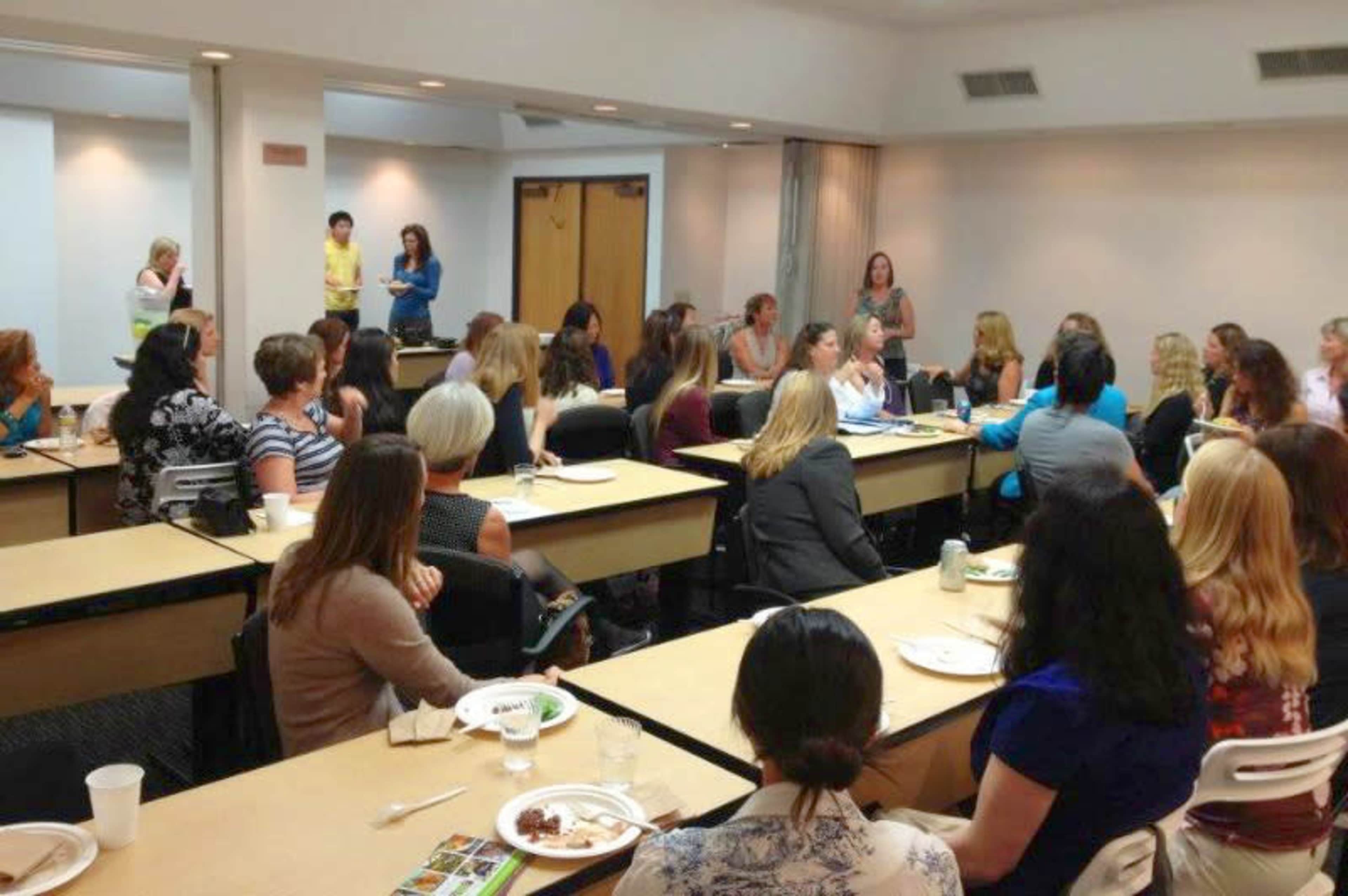 A group of women sit at tables in a conference room, engaged in discussion during a meeting.