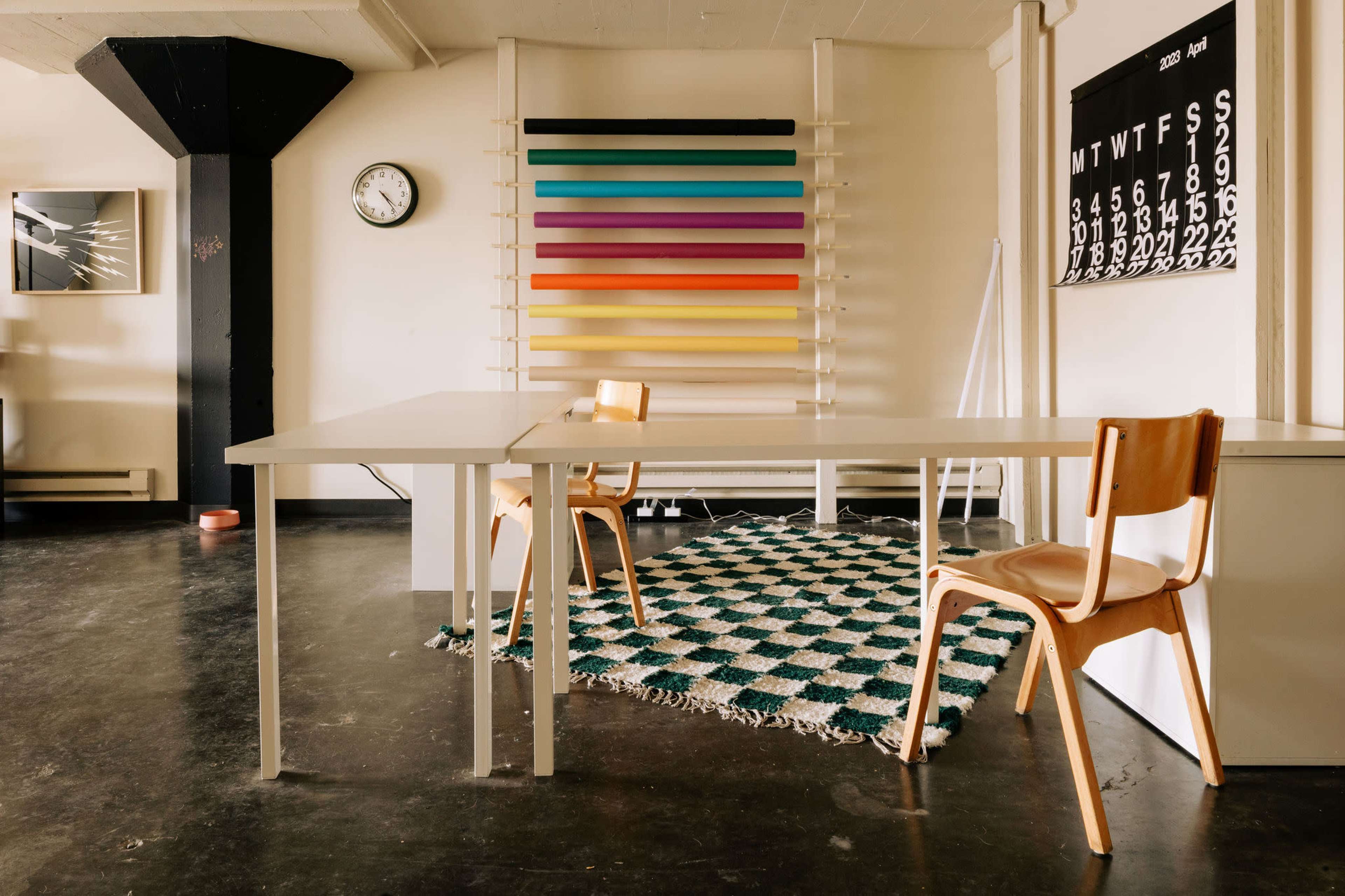 A minimalist interior with two wooden chairs at a white table, a patterned rug on the floor, and a colorful vertical display mounted on the wall.