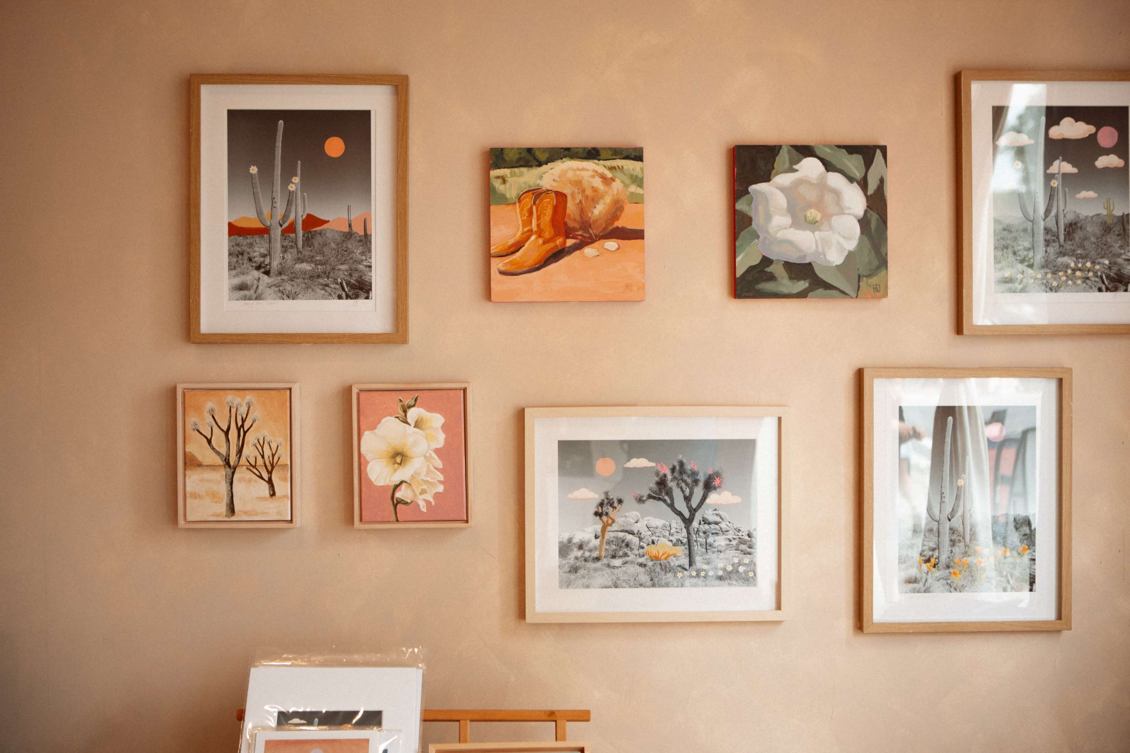 The image features a wall adorned with various framed paintings, showcasing desert landscapes, flowers, and a loaf of bread.
