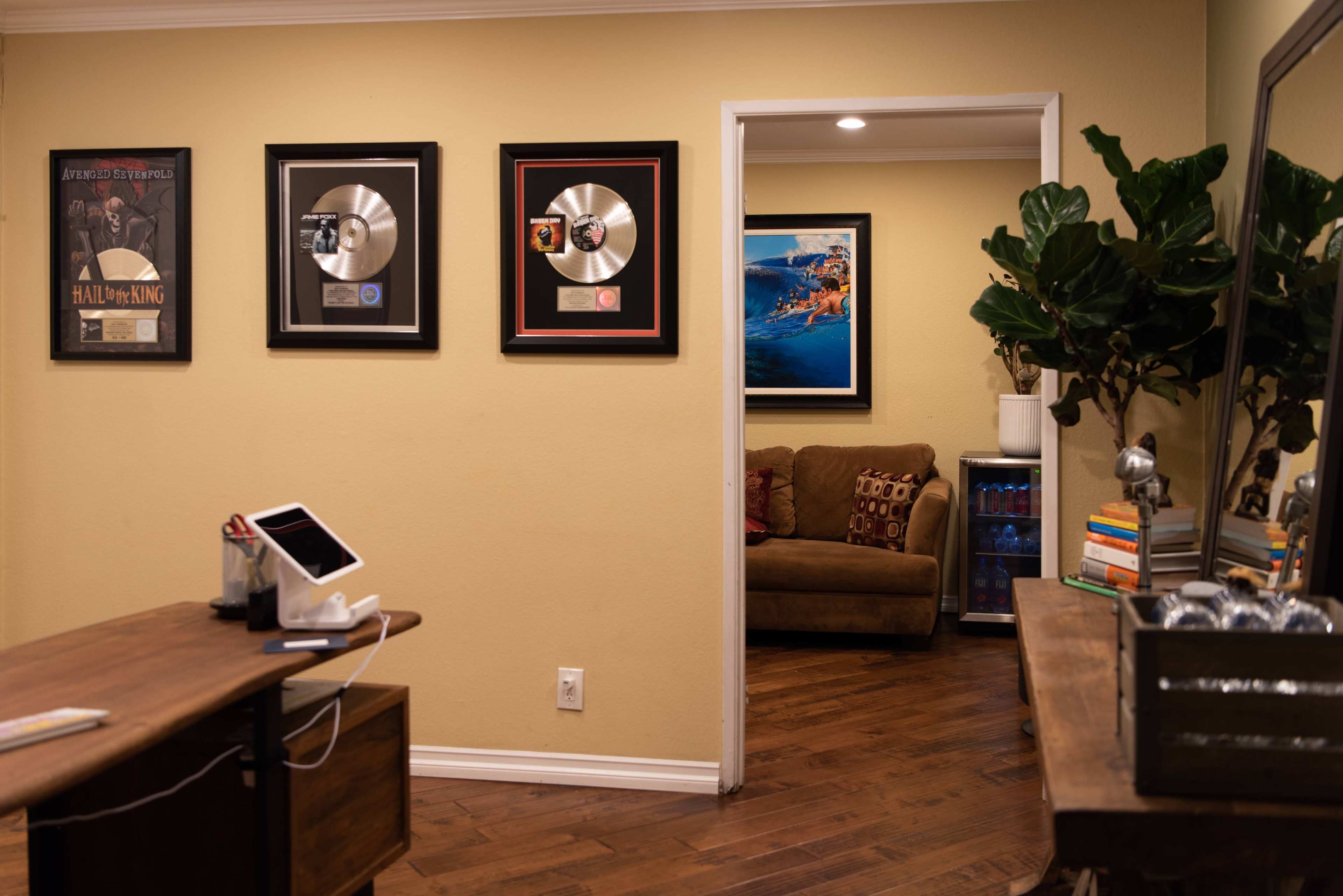 The image shows a cozy home office featuring gold and platinum record plaques on the wall, a wooden desk with a tablet stand, and a doorway leading to a living area with a couch and a plant nearby.
