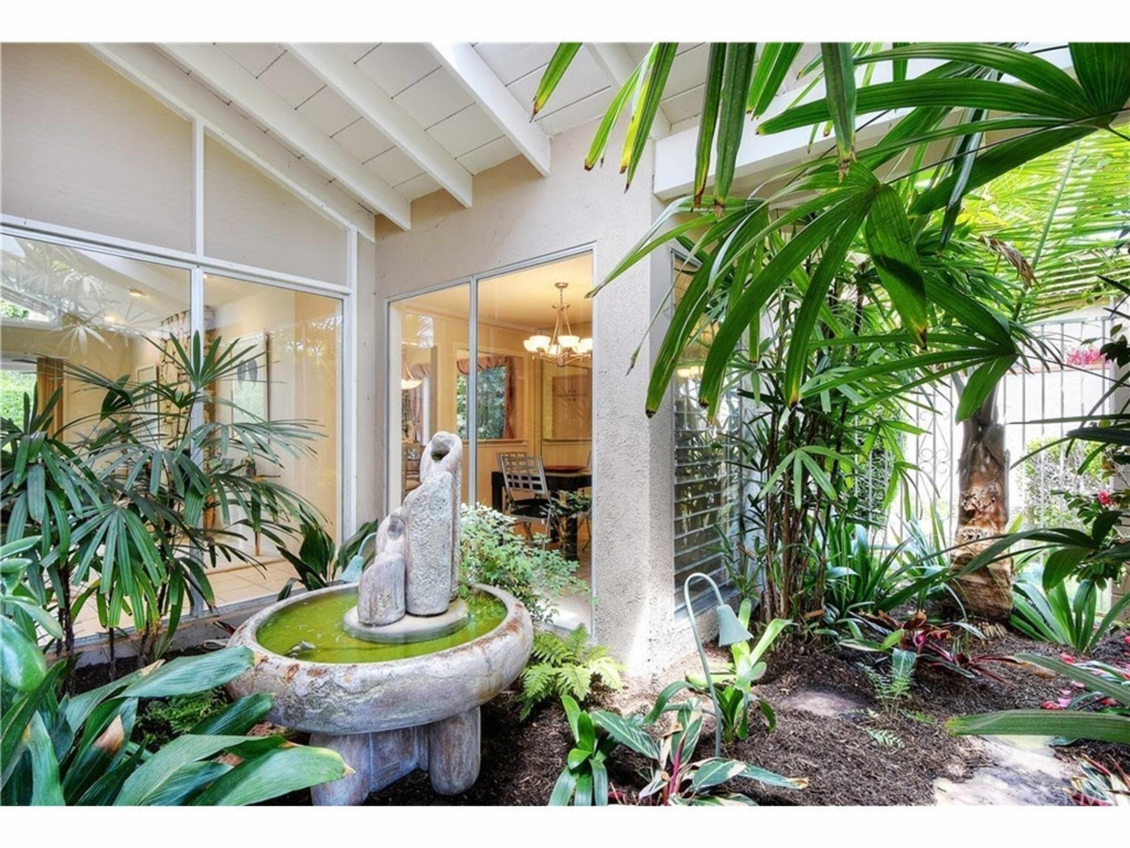 A stone fountain is centered in a lush garden surrounded by tropical plants, with a view of an adjacent room through large glass windows.