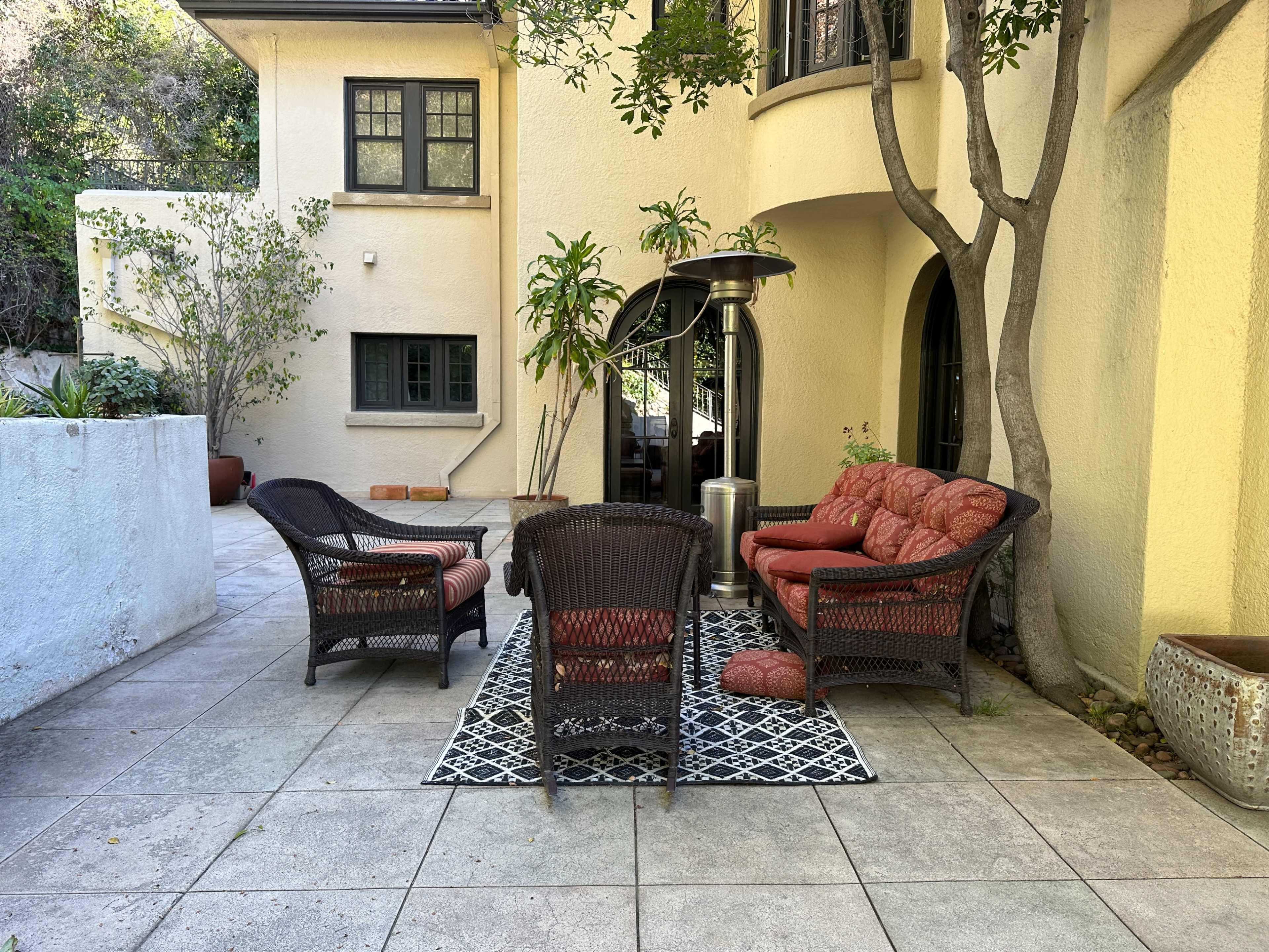 A patio area features a wicker seating arrangement with cushions, a patterned rug, and a patio heater, surrounded by greenery and a yellow stucco wall.