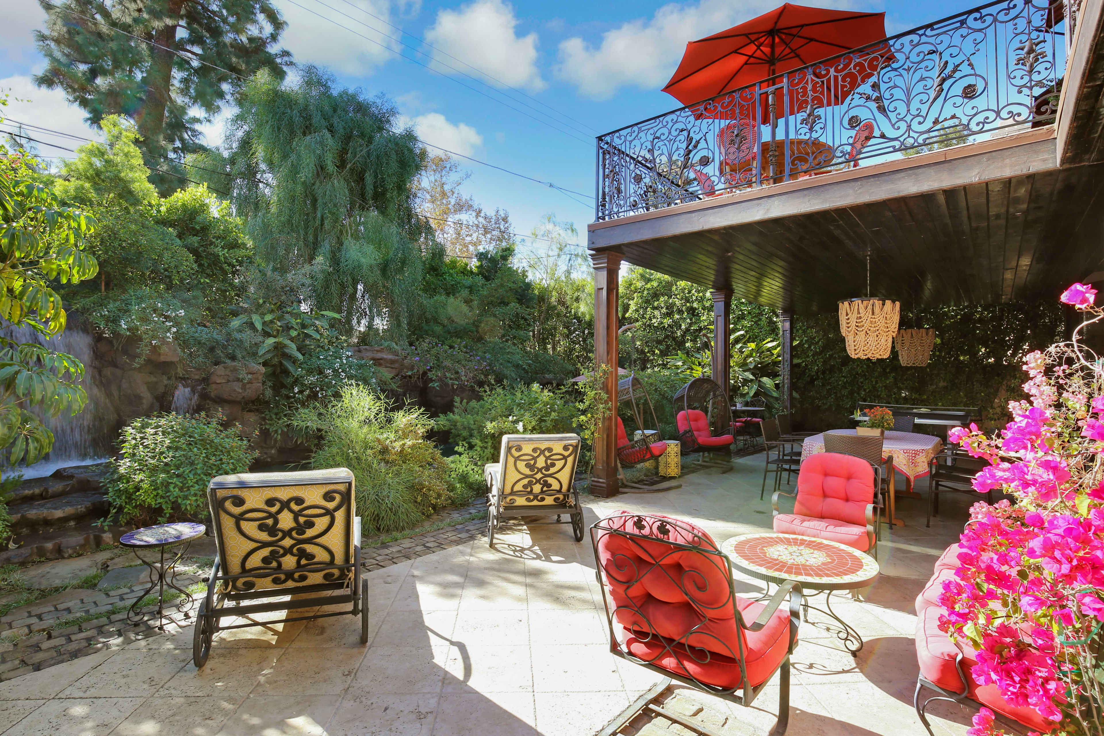 A backyard patio area with wrought iron furniture, a table, and a second-story balcony surrounded by lush greenery and a small waterfall.