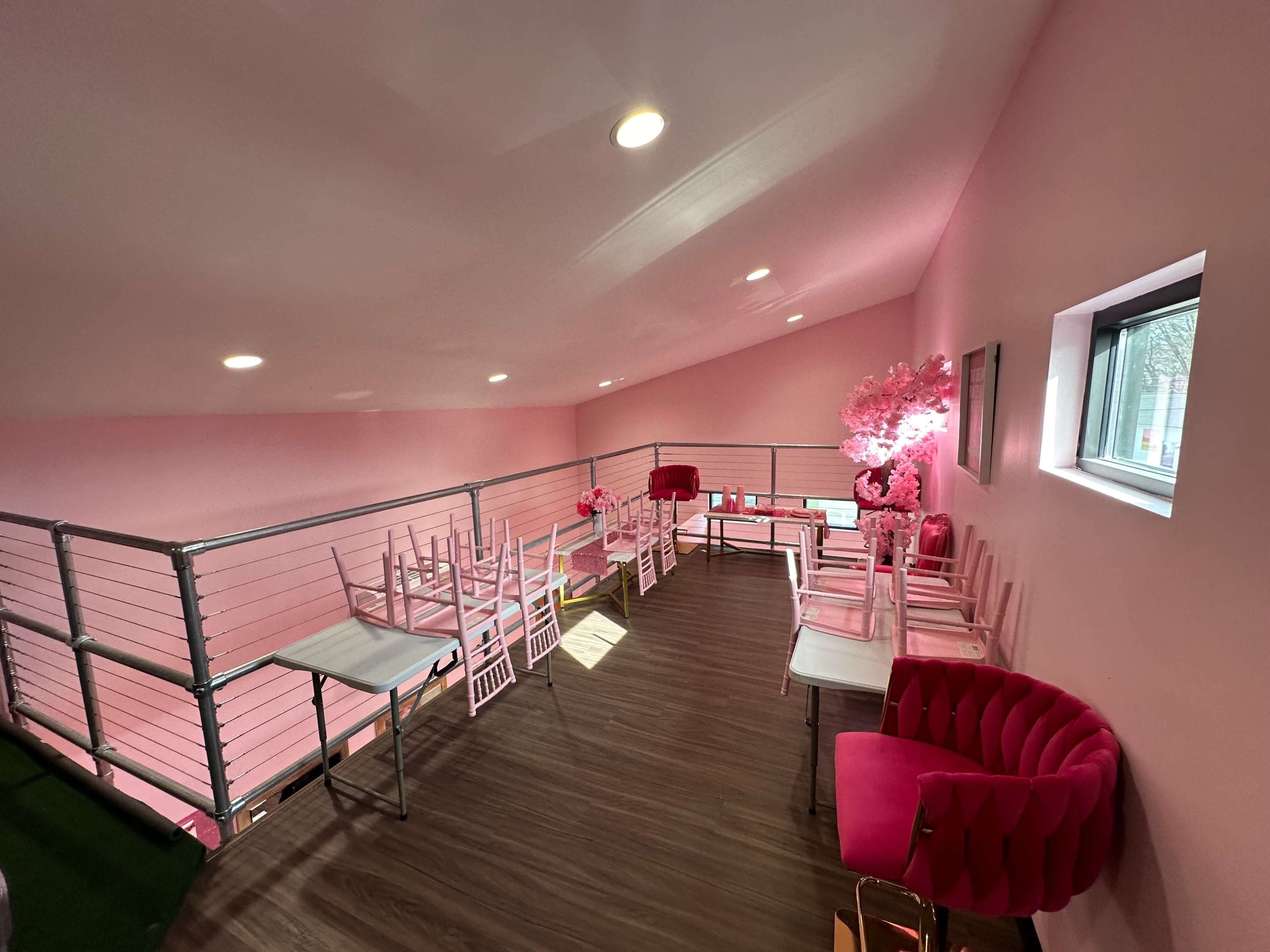 The image shows a bright room with pink walls, several white tables and chairs, and a pink accent chair near a window.