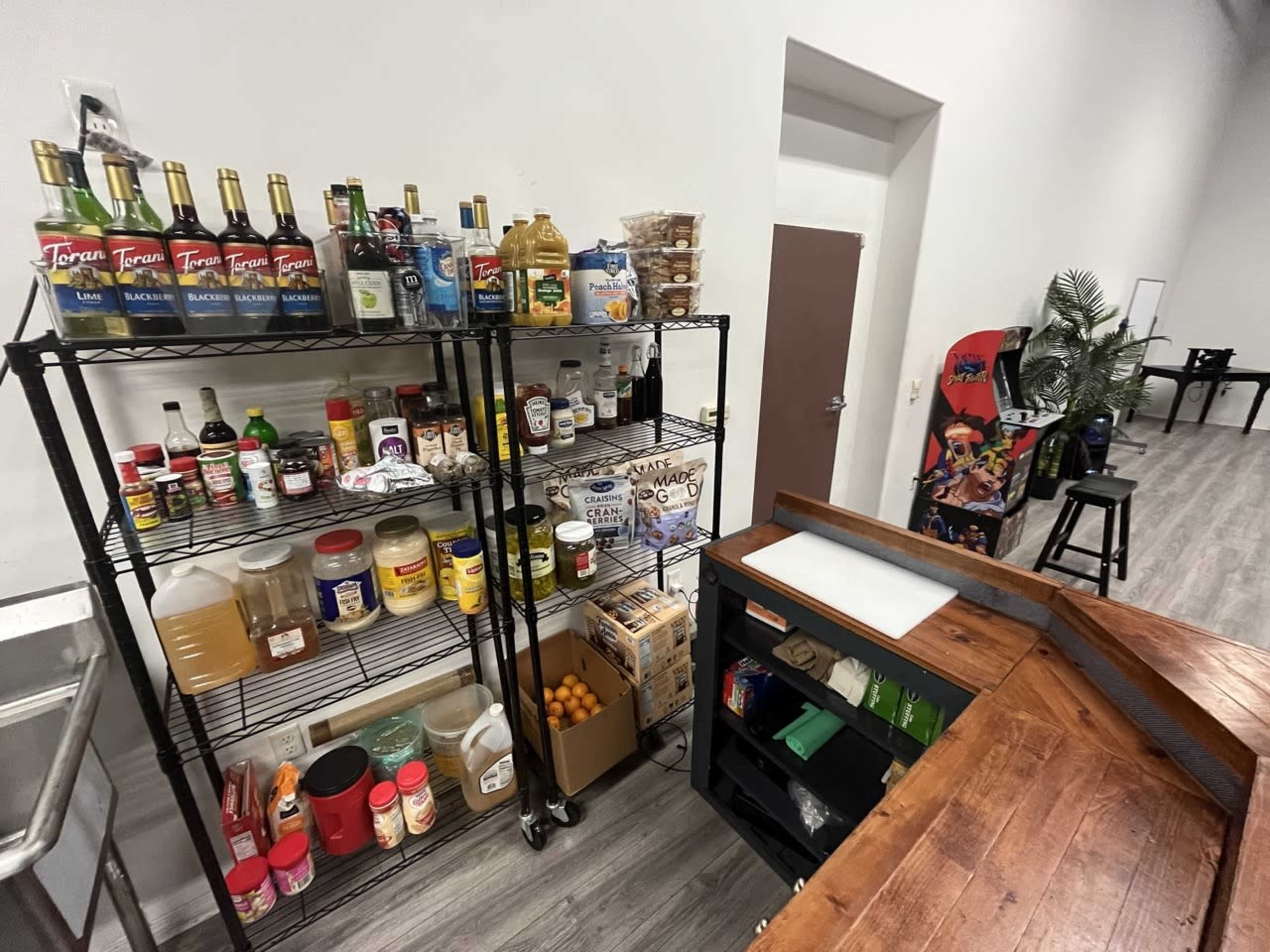 A well-organized kitchen storage area with shelves containing various condiments, snacks, and ingredients next to a wooden counter and a small table in the background.