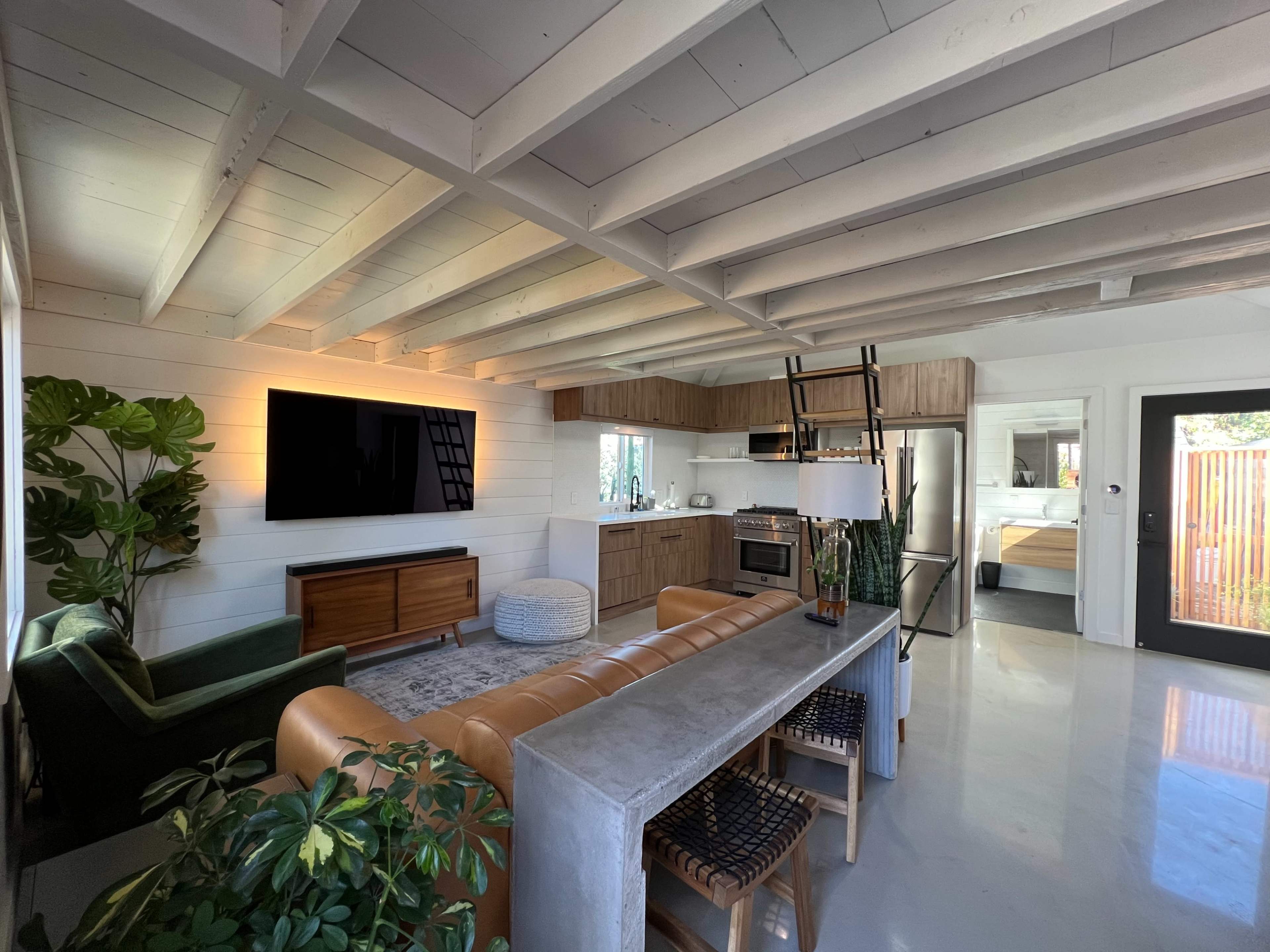 The image depicts a modern open-concept living space featuring a kitchen, living area, and dining area with exposed beams and contemporary furnishings.