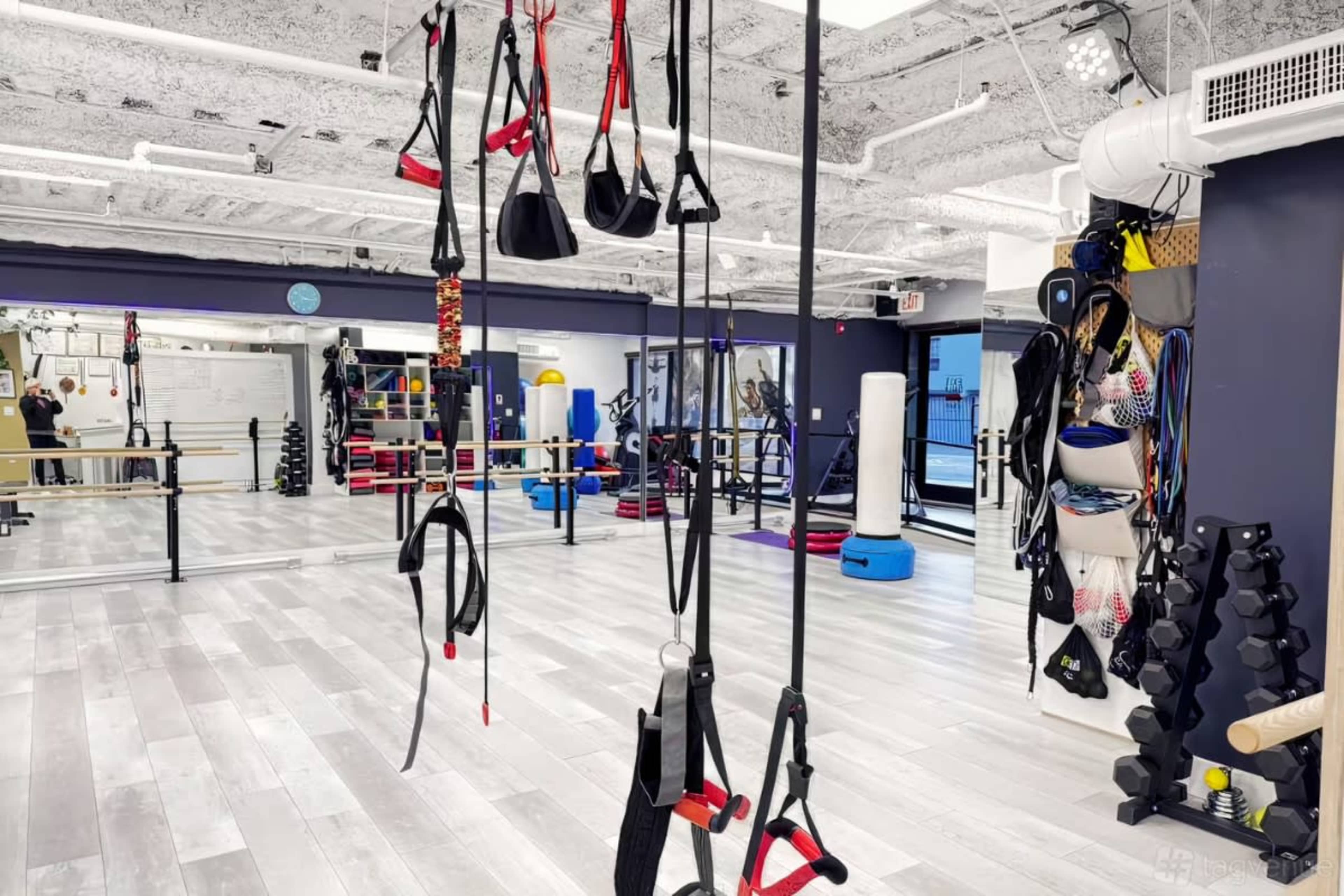 The image shows a well-lit fitness studio equipped with various exercise equipment, including resistance bands, weights, and a ballet bar.
