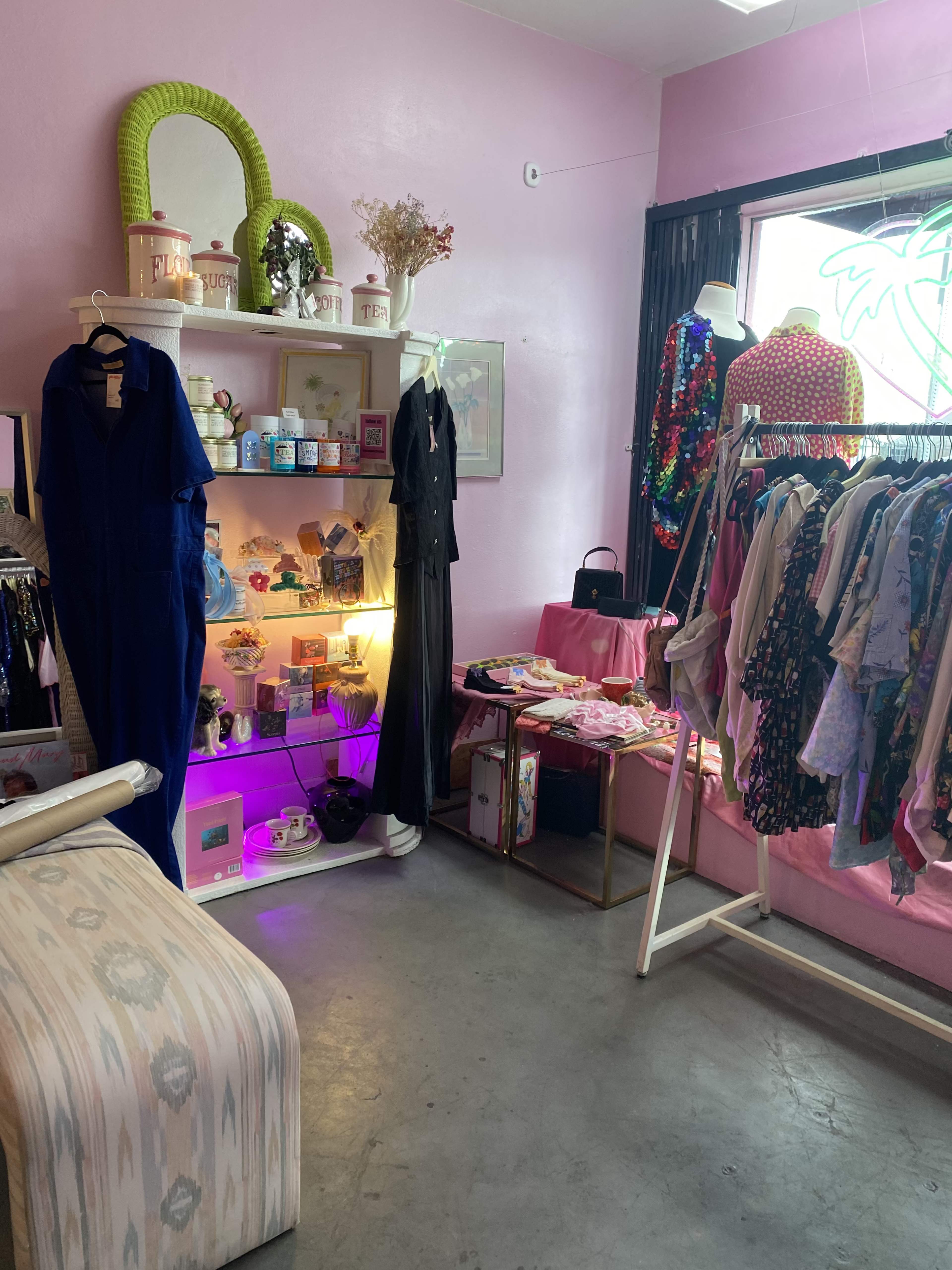 A brightly decorated boutique with clothing racks filled with various garments, a shelf displaying accessories and decor, and a pink wall in the background.