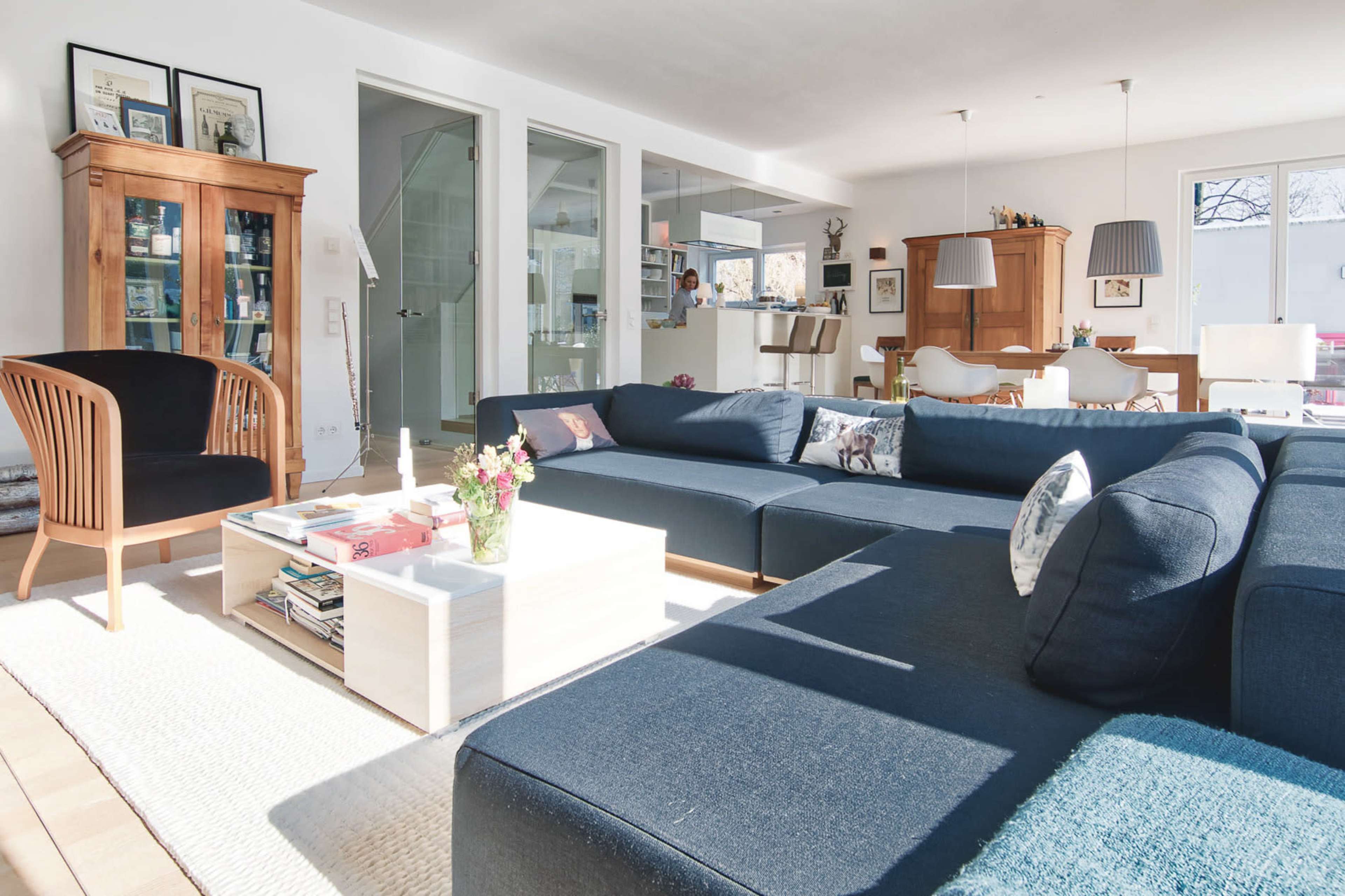 The image shows a modern living room with a large sectional sofa, a coffee table, a wooden chair, and a dining area visible in the background.